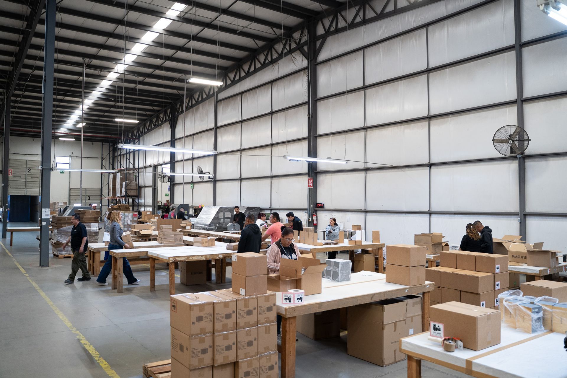 People packing boxes in a large warehouse with tables and stacks of cardboard boxes