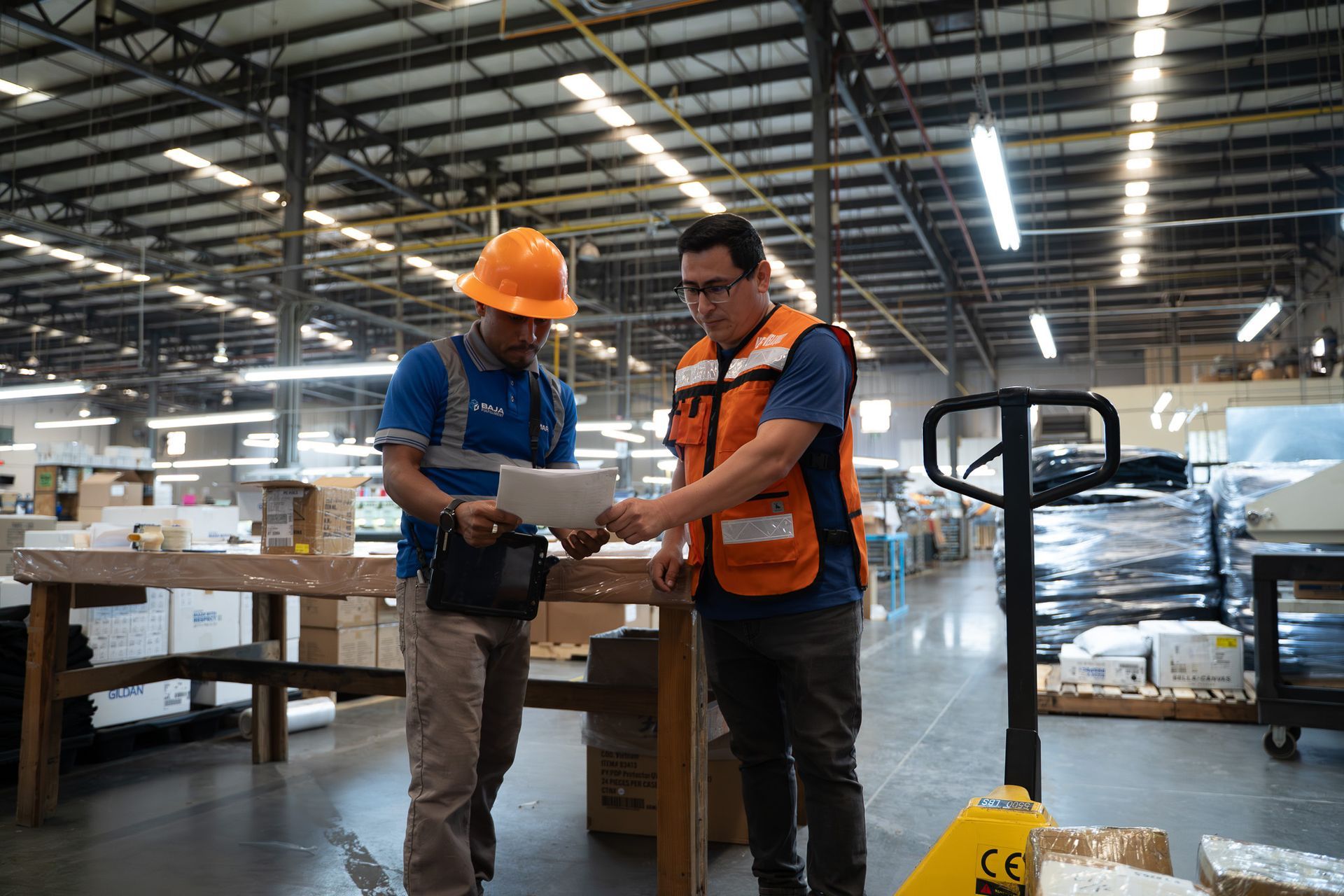 Two workers reviewing a document in a warehouse, one wearing a hard hat and orange safety vest.