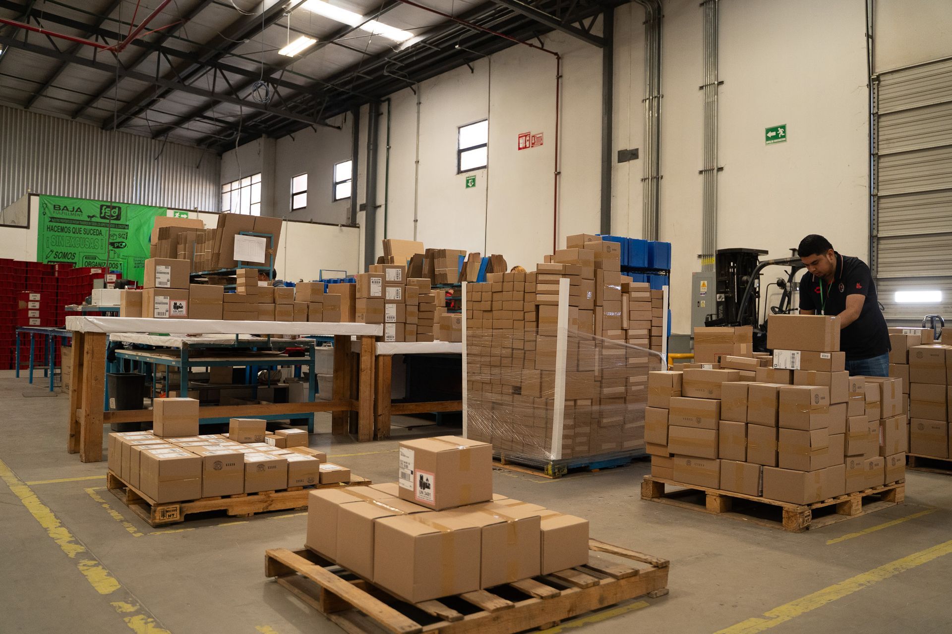 Warehouse with stacked cardboard boxes on pallets and a worker using a forklift near the loading area