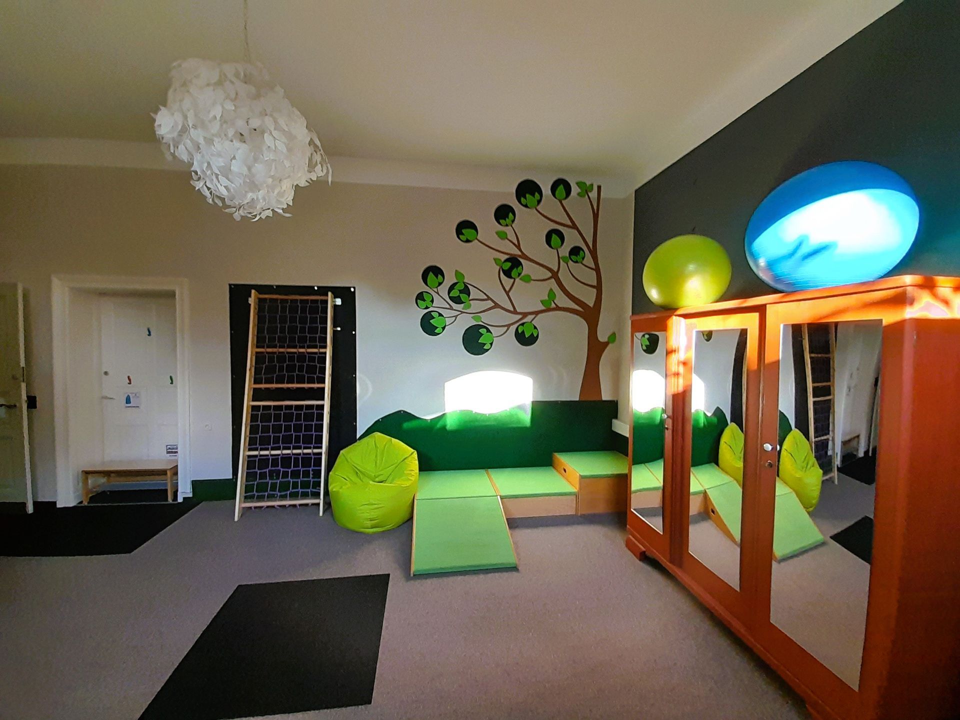 Spielzimmer mit grünen und braunen Möbeln, einem Baumwandbild und bunten Bällen auf einem Schrank.