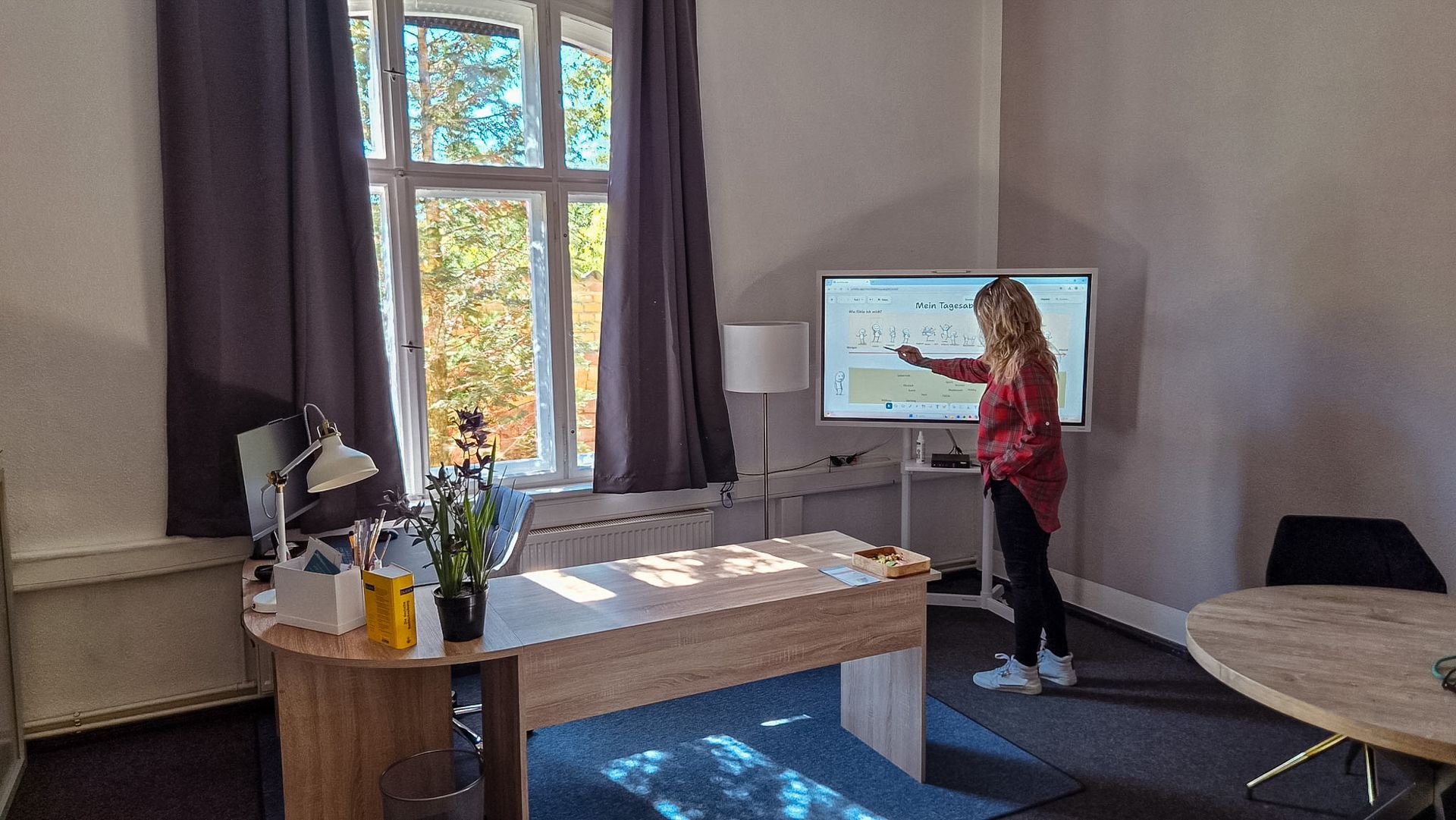 Eine Frau hält einen Vortrag neben einem Fernseher in einer hellen Bürolounge mit einem großen Fenster und einem Tisch.