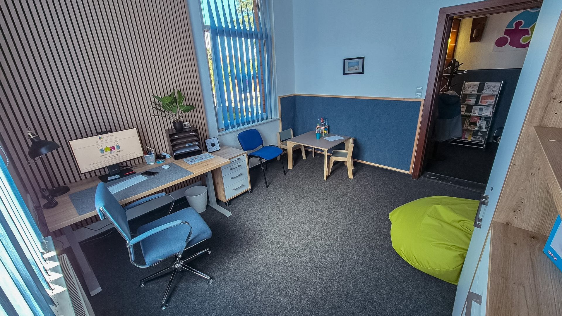 Kleines Büro mit einem Schreibtisch, Stühlen, einem Laptop und einem grünen Sitzsack in einem blau-weißen Raum