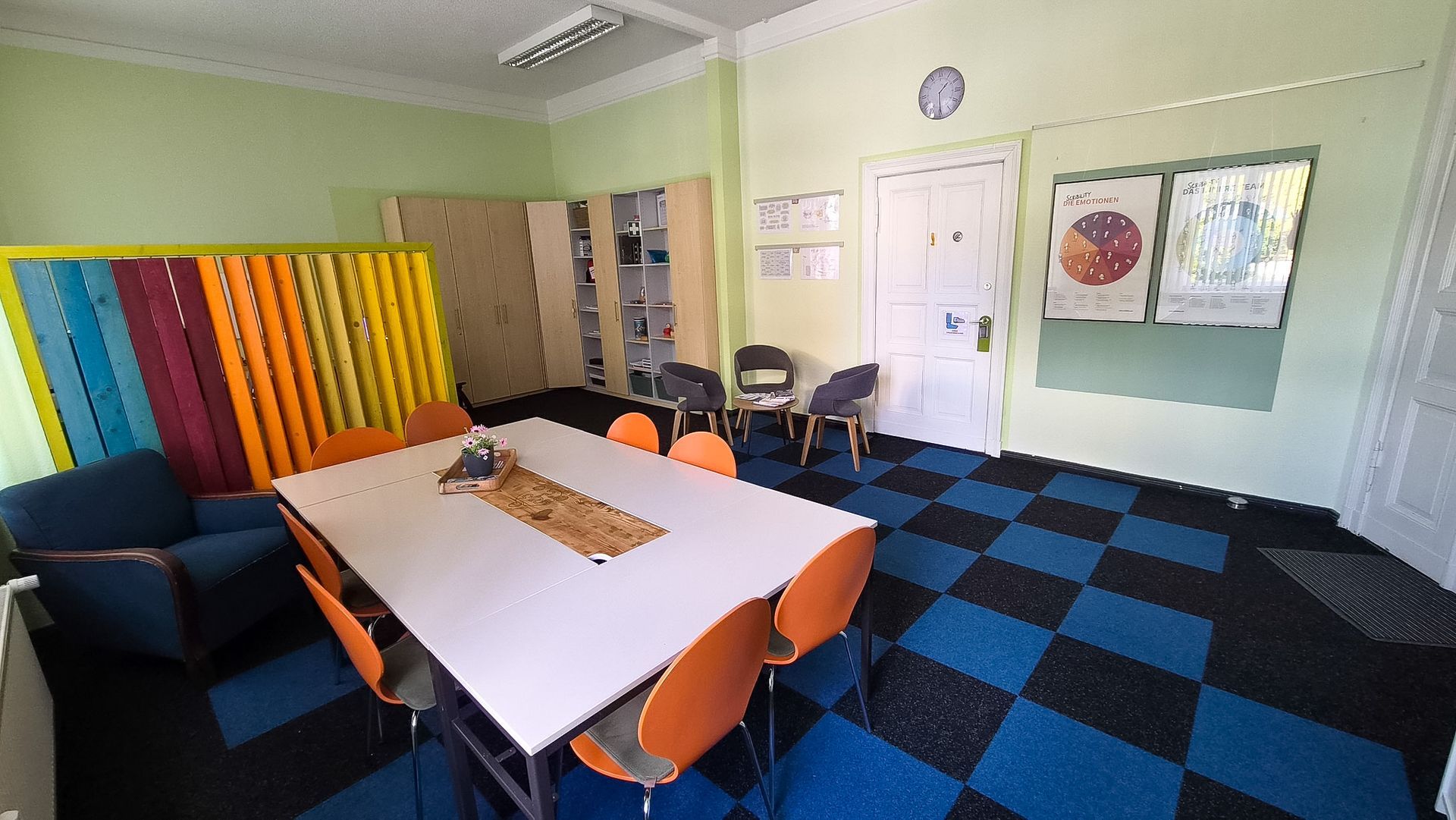 Heller Klassenraum mit blau-schwarz kariertem Boden, orangefarbenen Tischen, Stühlen, bunten Wandpaneelen und Whiteboards.