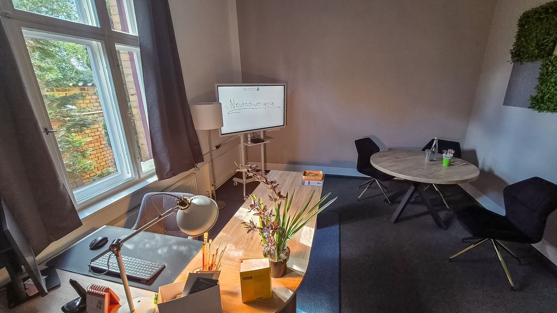 Helles Büro mit einem Schreibtisch am Fenster, einem Whiteboard, einem Tisch, Stühlen und Topfpflanzen auf dem Teppichboden.