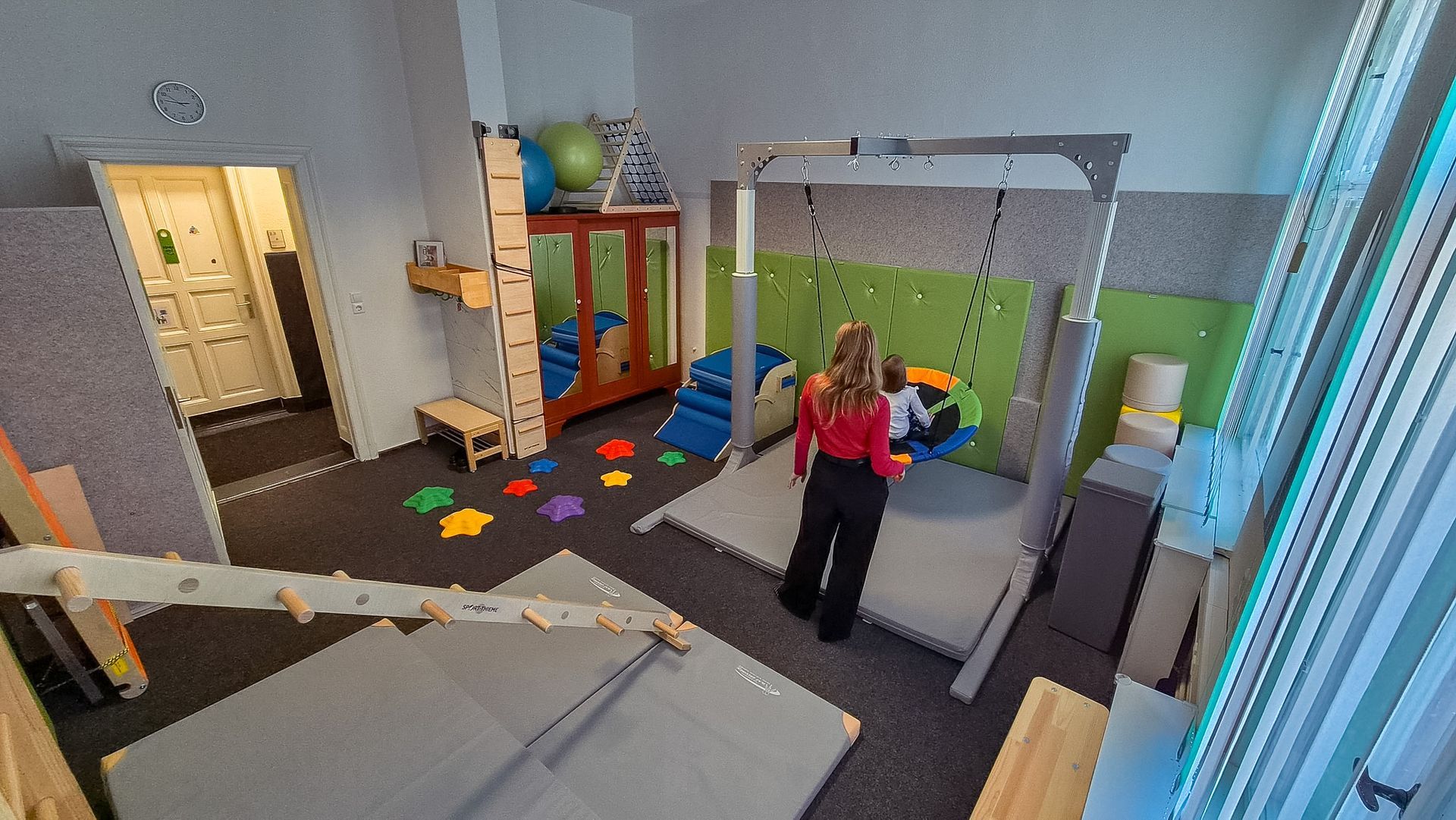 Ein Kind im roten T-Shirt sitzt auf einem Trampolin in einem Spielzimmer mit Spielzeug und Regalen.
