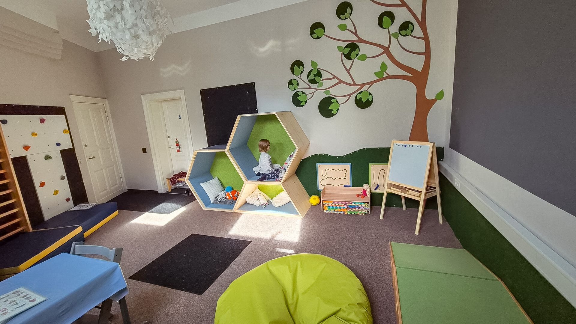 Gemütliches Spielzimmer für Kinder mit sechseckigen Sitzgelegenheiten, einem Wandbild mit einem Baum, Spielzeug und einem kleinen Tisch am Fenster