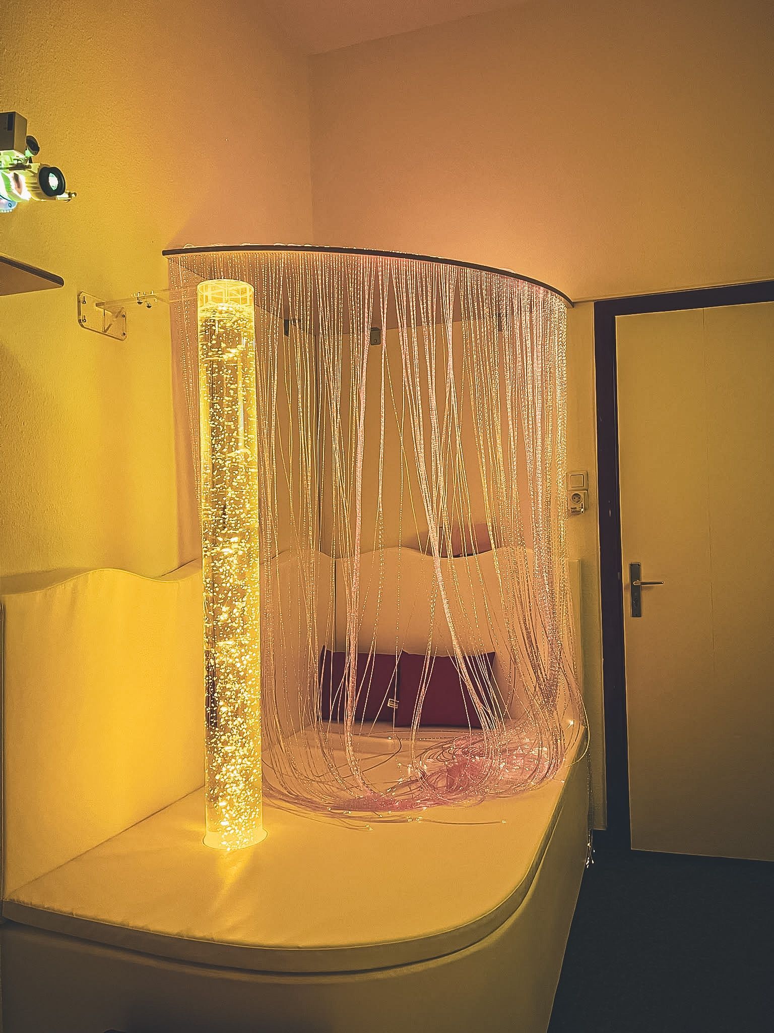 Gebogene Bühne mit schimmerndem goldenem Vorhang und einem Stuhl, in warmes gelbes Licht getaucht, neben einer offenen Tür