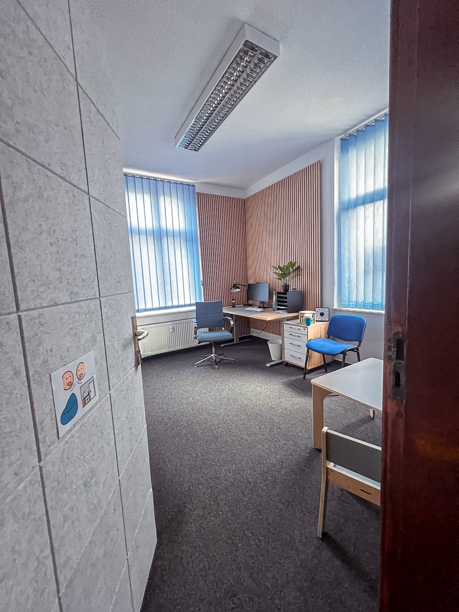 Kleines Büro mit blauen Stühlen, einem Schreibtisch, hohen Fenstern und gestreiften Vorhängen.