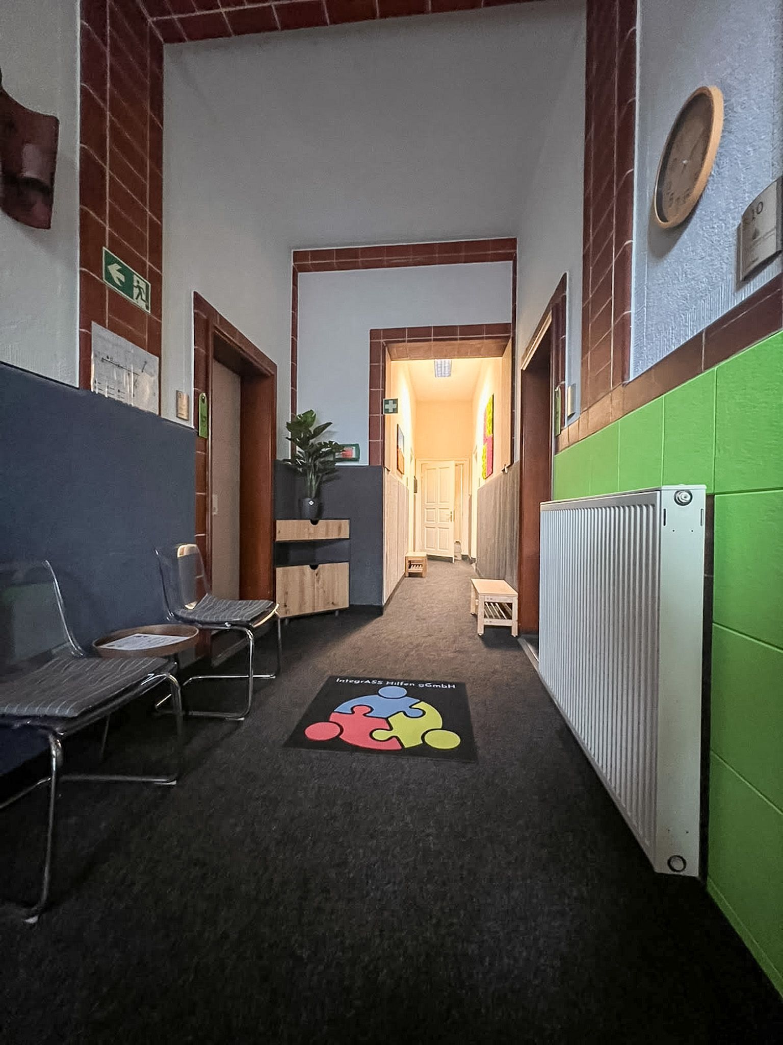 Schmaler Flur mit schwarzem Teppich, schwarzer Sitzbank, grünen Wänden und einem hell erleuchteten Durchgang am Ende