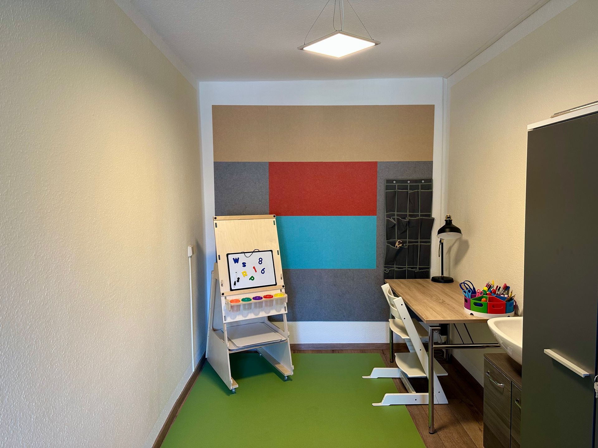 Schmaler Raum mit farbenfrohen Wandpaneelen, einem Schreibtisch, Stühlen und einer kleinen Staffelei am hinteren Ende.