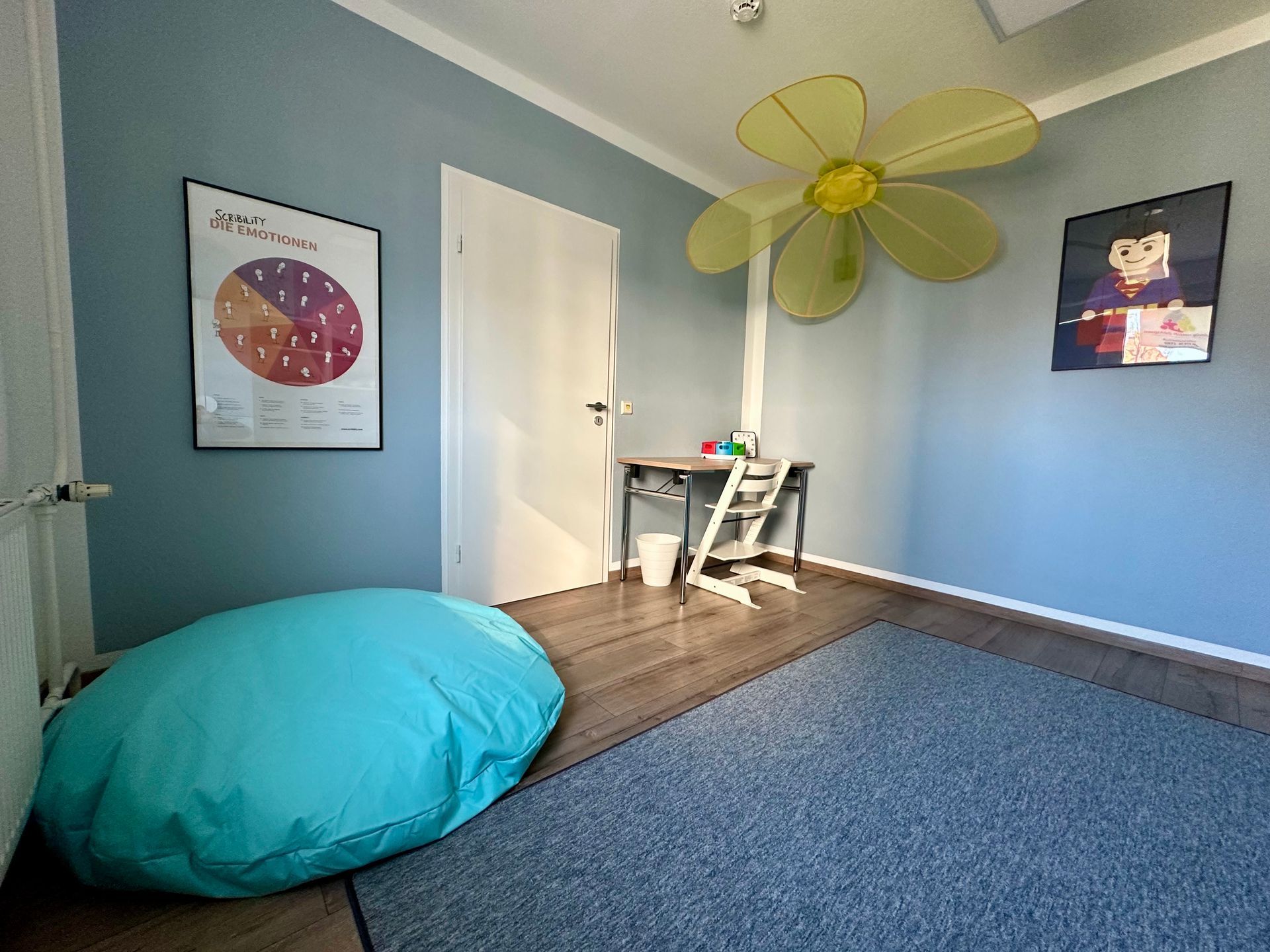 Zimmer mit blauen Wänden, Sitzsack, Poster, blumenförmigem Deckenventilator, Schreibtisch und Tür.
