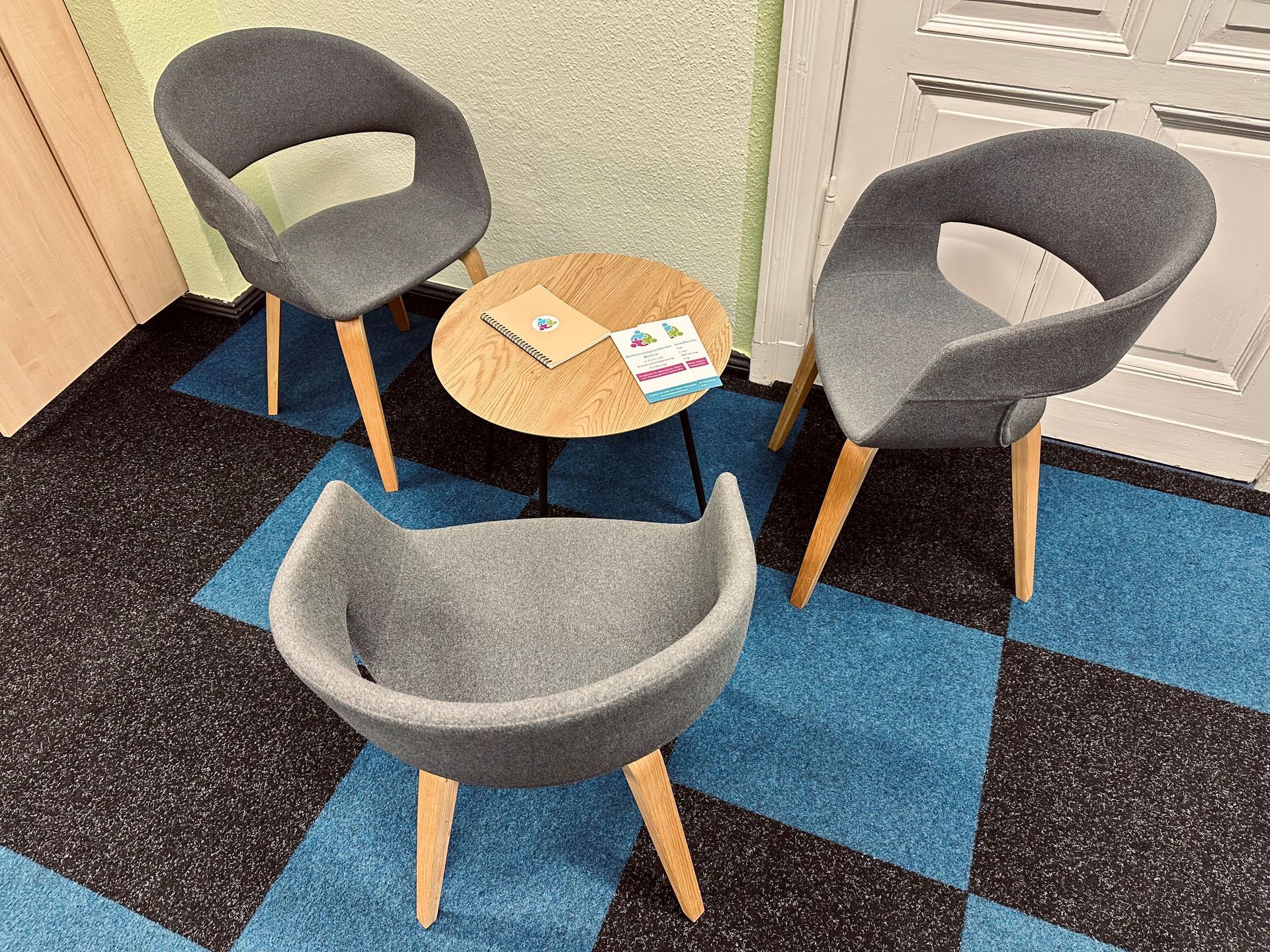 Drei graue Stühle umgeben einen kleinen Holztisch. Die Stühle haben Holzbeine und stehen auf einem blau-schwarz gemusterten Boden.