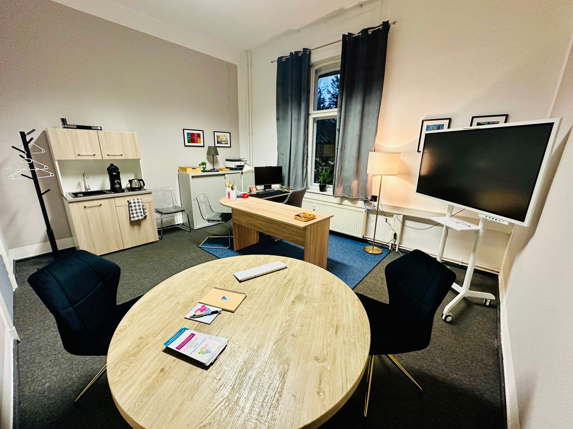 Büroeinrichtung: runder Tisch mit Stühlen, Schreibtisch mit Monitor, kleine Küchenzeile, Fenster mit dunklen Vorhängen.