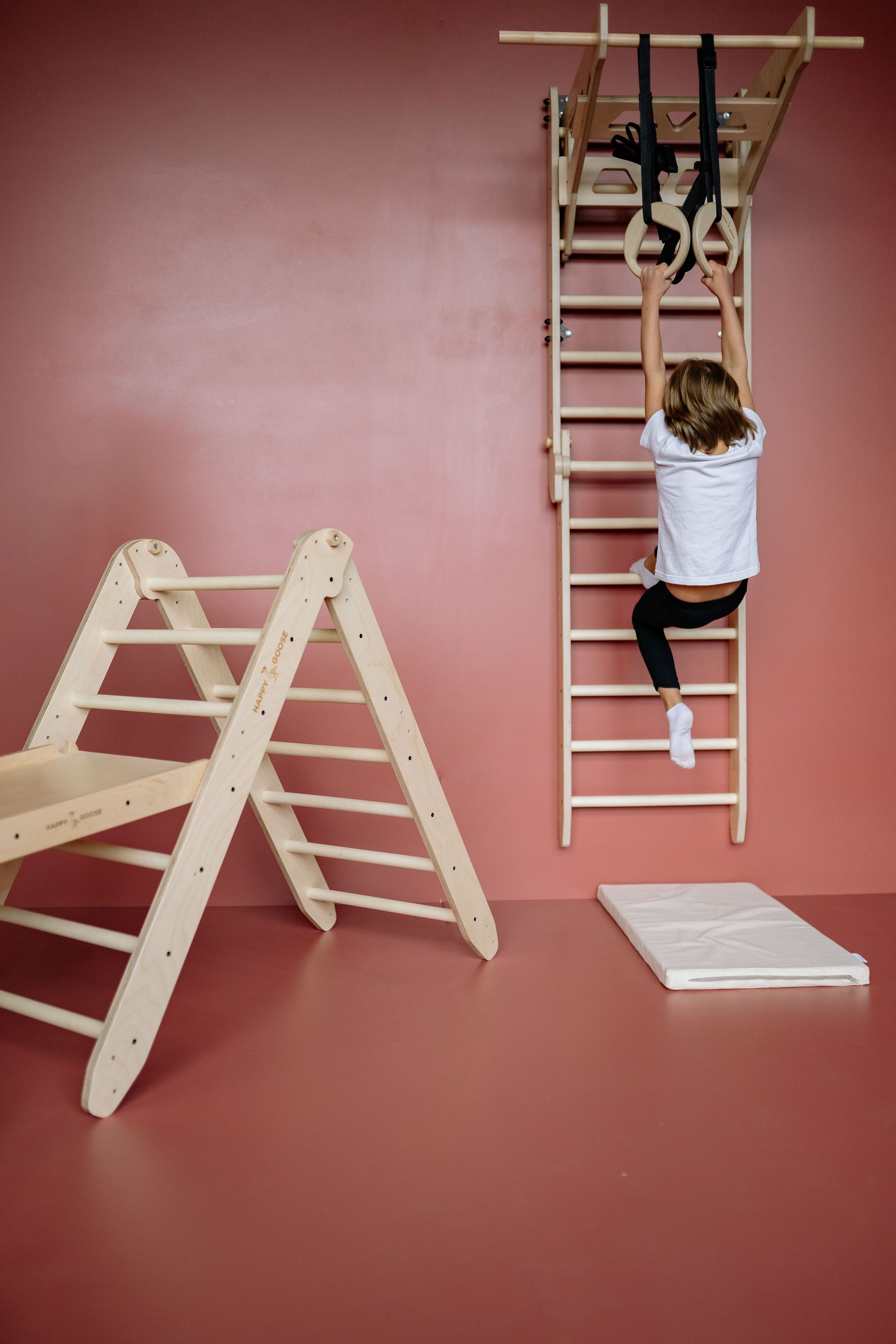 Eine Person klettert neben einer A-Leiter eine Leiter hinauf. Daneben liegt ein Stapel Bretter auf einem pinkfarbenen Boden und vor einer pinkfarbenen Wand.