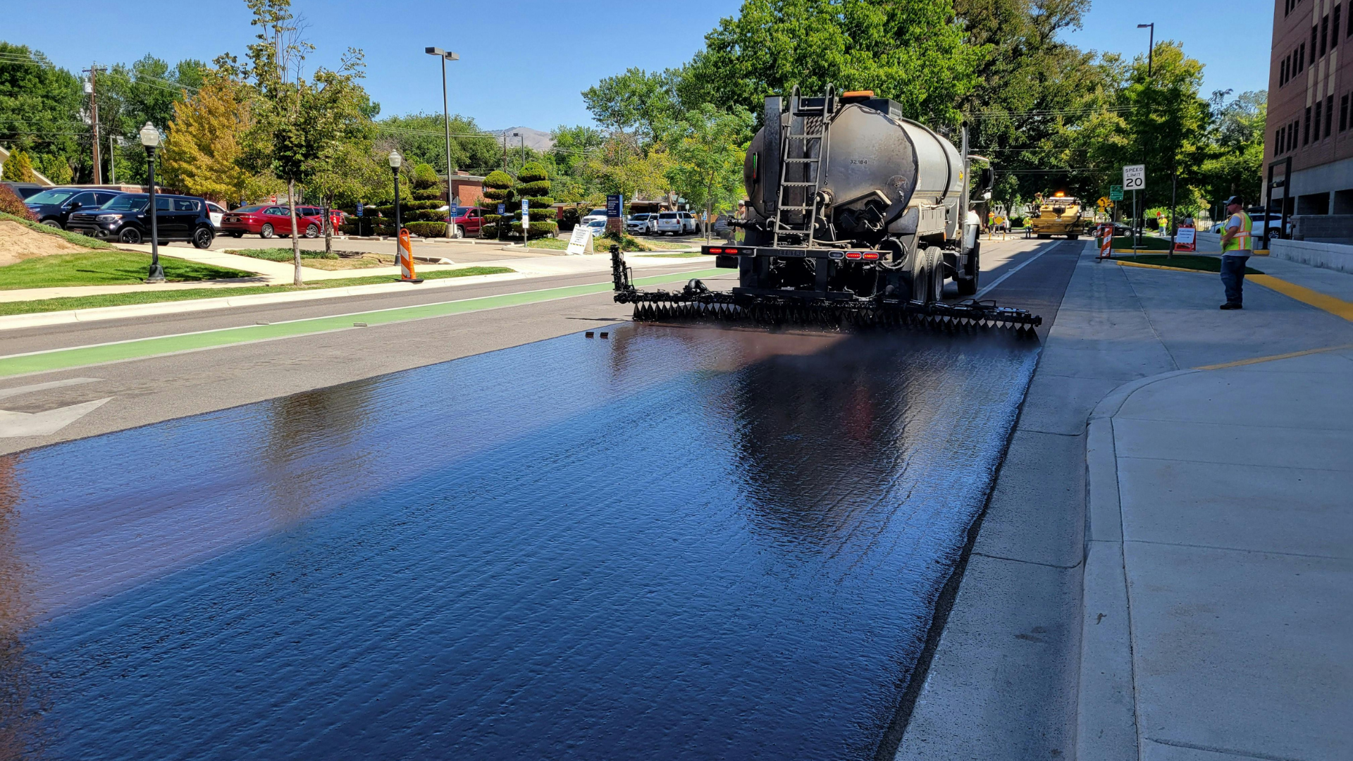 Water truck spraying asphalt on a sunny suburban street with trees and parked cars nearby