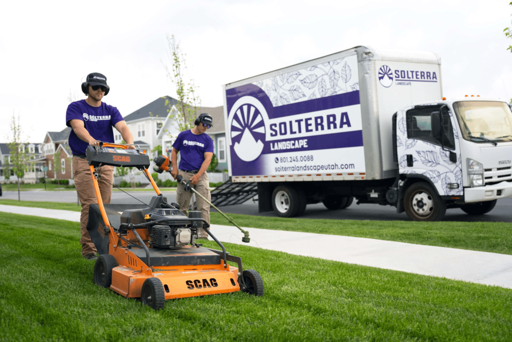 Two men mowing a lawn with a truck that says Solterra Landscape parked in the background.