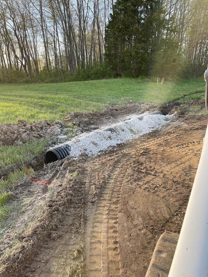 Dirt construction site with a culvert pipe, muddy trench, and excavator tracks beside a grassy field