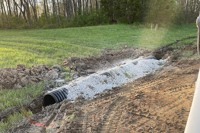 Black drainage pipe discharging water into a muddy ditch beside a grassy field