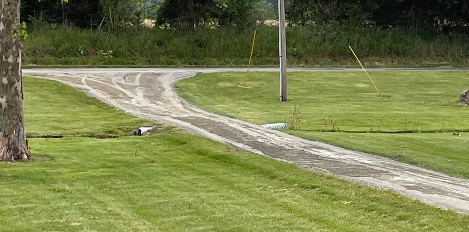 Curving gravel driveway through a green lawn beside a road, with a utility pole and trees nearby