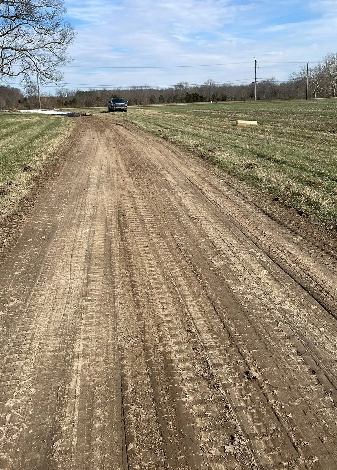 Dirt road through open fields under a partly cloudy sky, with a car in the distance.