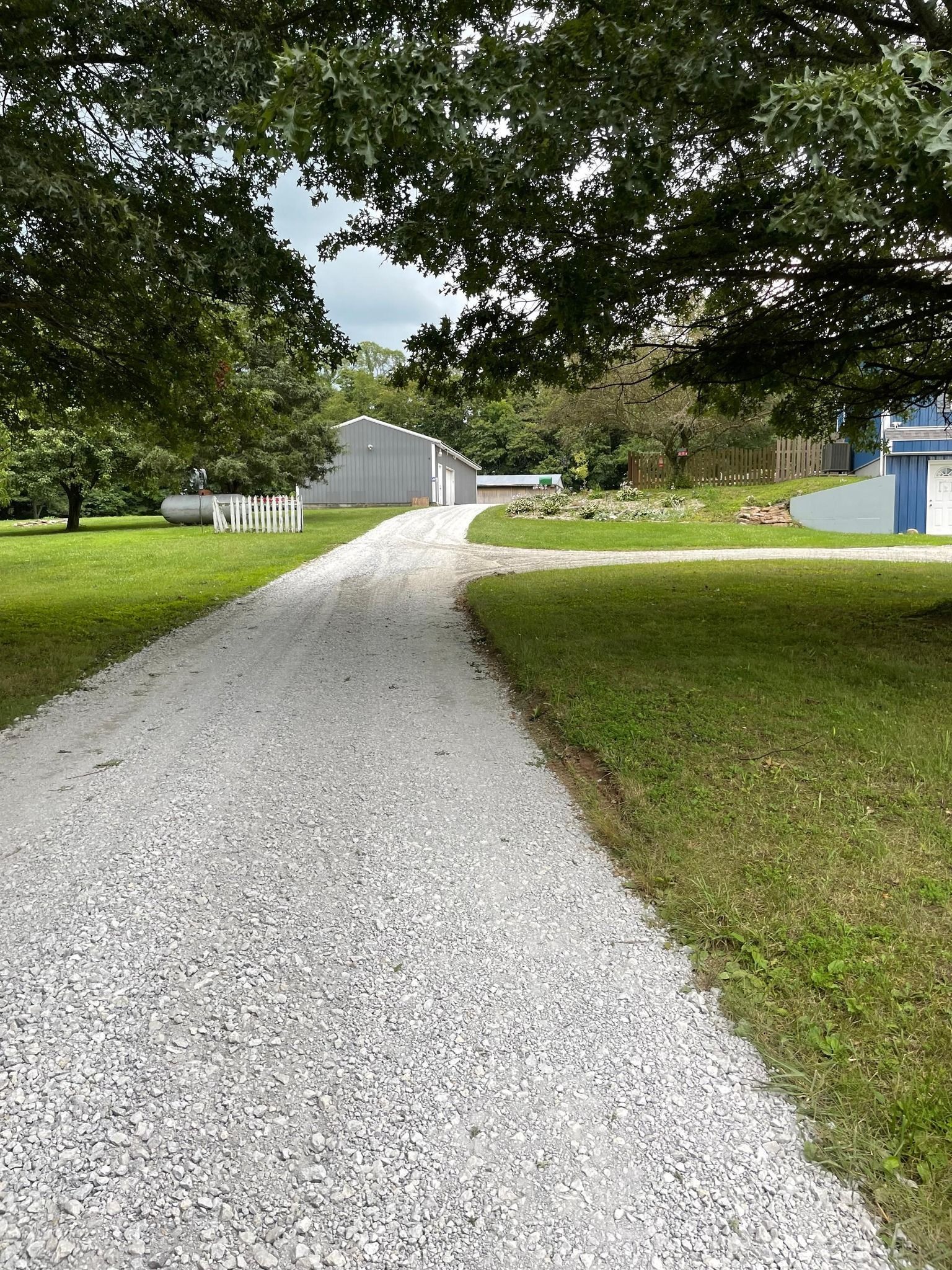 Gravel driveway curving through a grassy yard with trees and a barn-like building in the distance