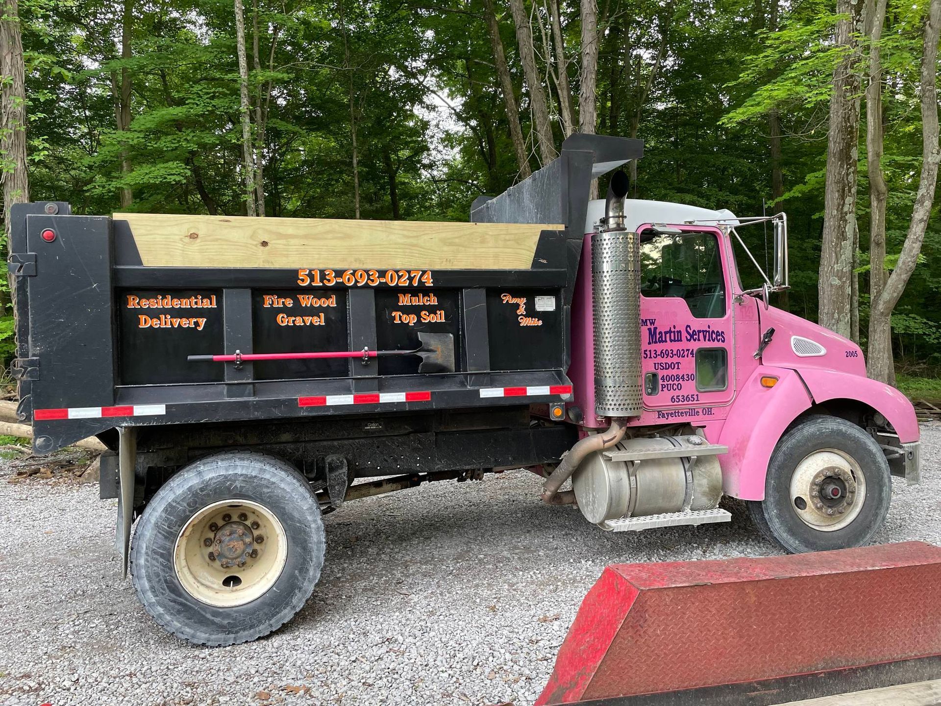 Pink dump truck parked on gravel beside red barrier in a wooded area