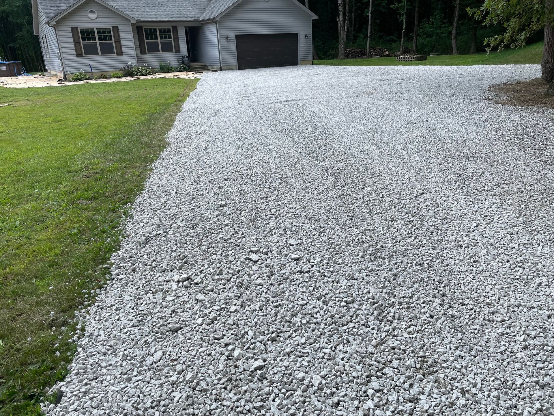 Gravel driveway leading to a small house and garage, with green lawn and trees beside it