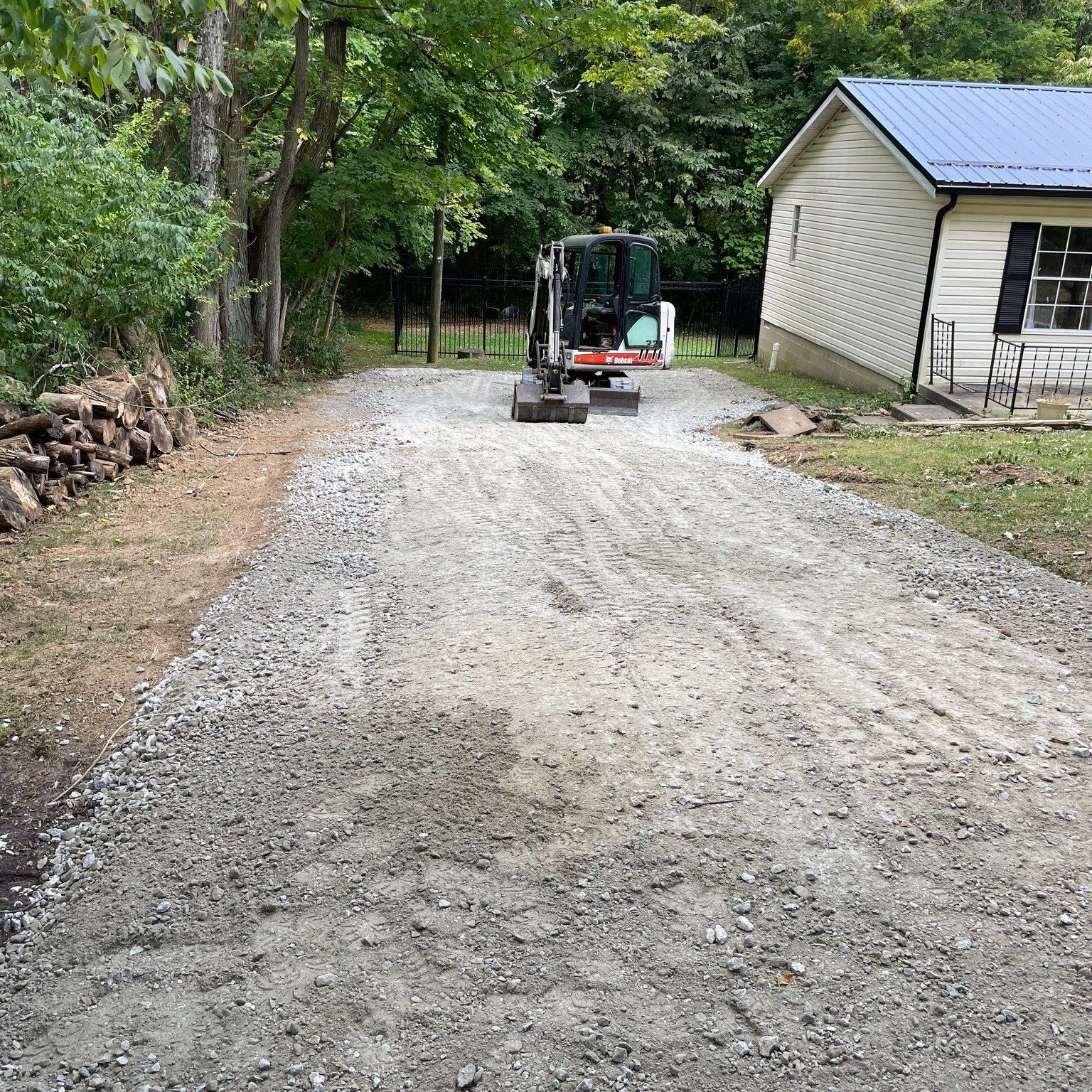 Gravel driveway under construction beside a beige house, with a compact excavator working near the end.