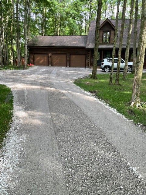Gravel driveway leading to a wooden house and garage in a wooded area, with a white vehicle parked outside