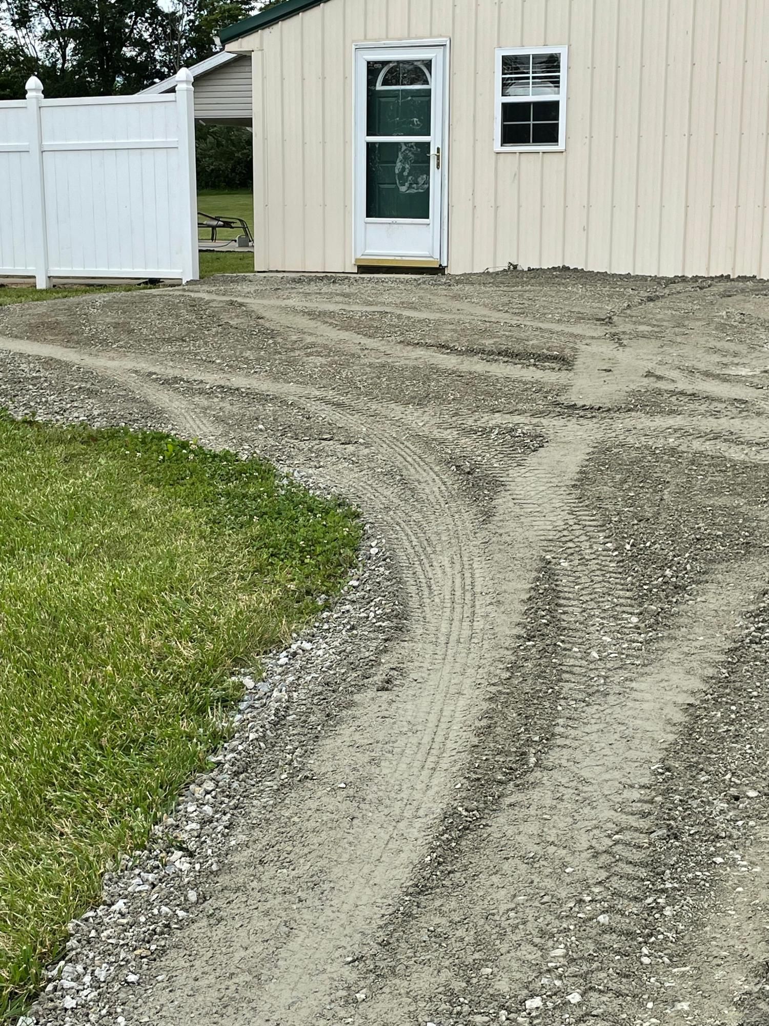 Gravel driveway curving past a beige metal building with a white fence and green grass.