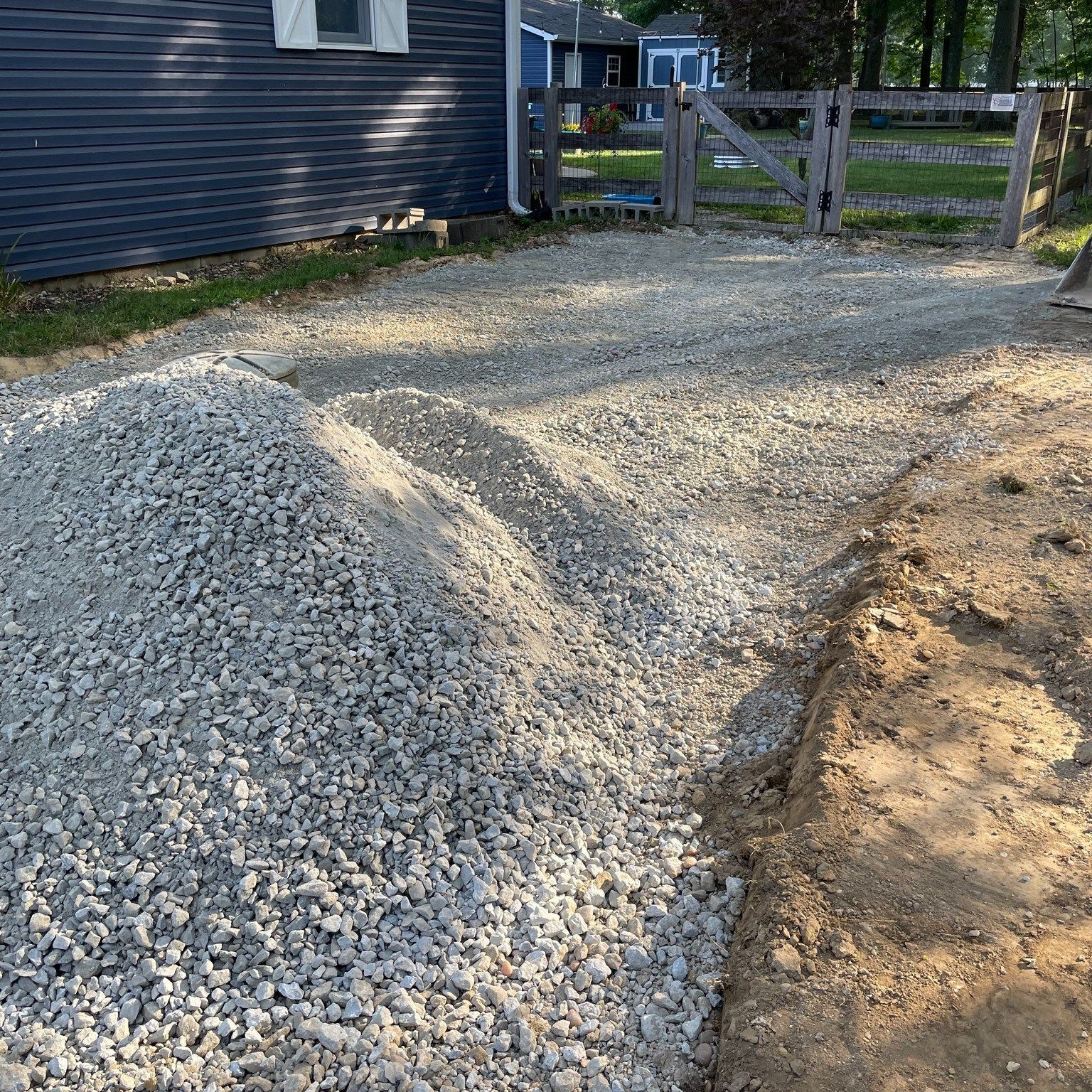 Gravel driveway under construction beside a house, with a pile of crushed stone near a wooden gate.