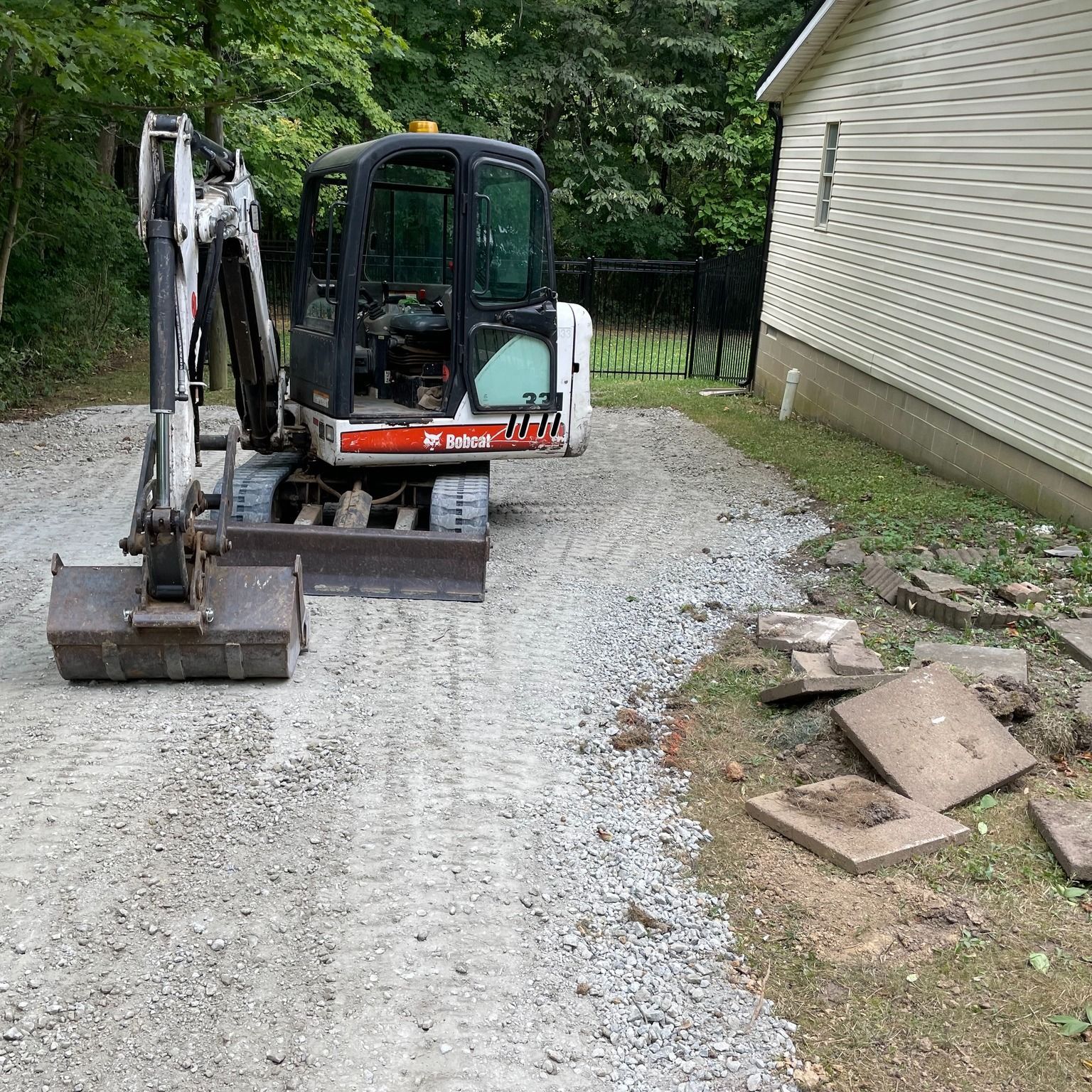 Mini excavator on a gravel driveway beside a house, with a pile of debris nearby.