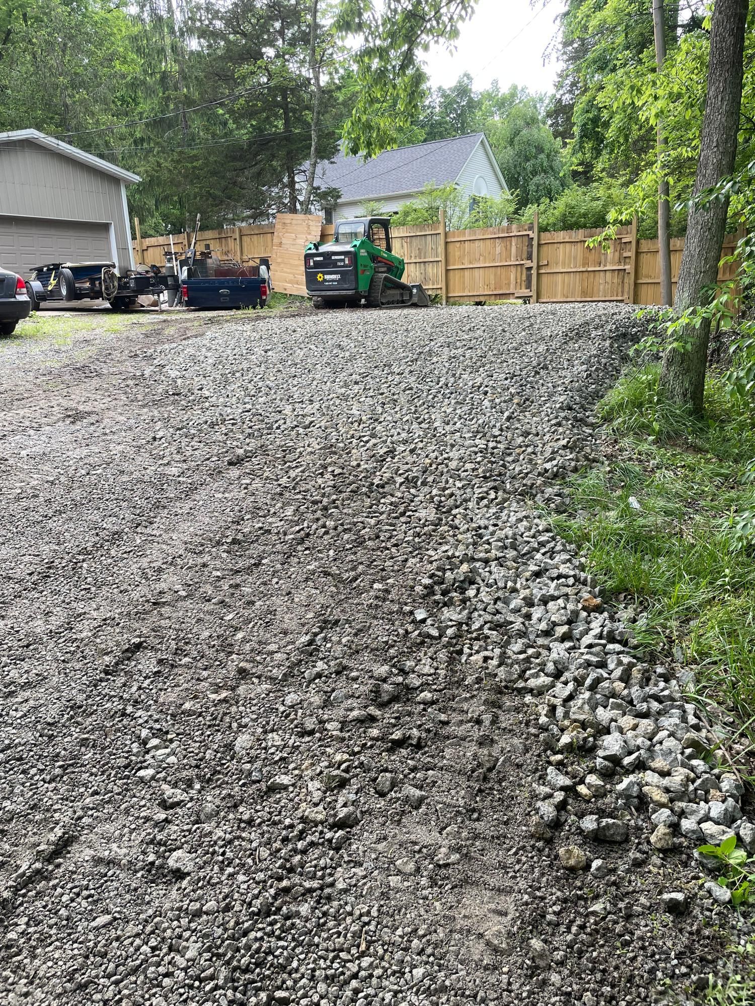 Gravel driveway under construction with a small green loader and wooden fence beside a house in a wooded yard