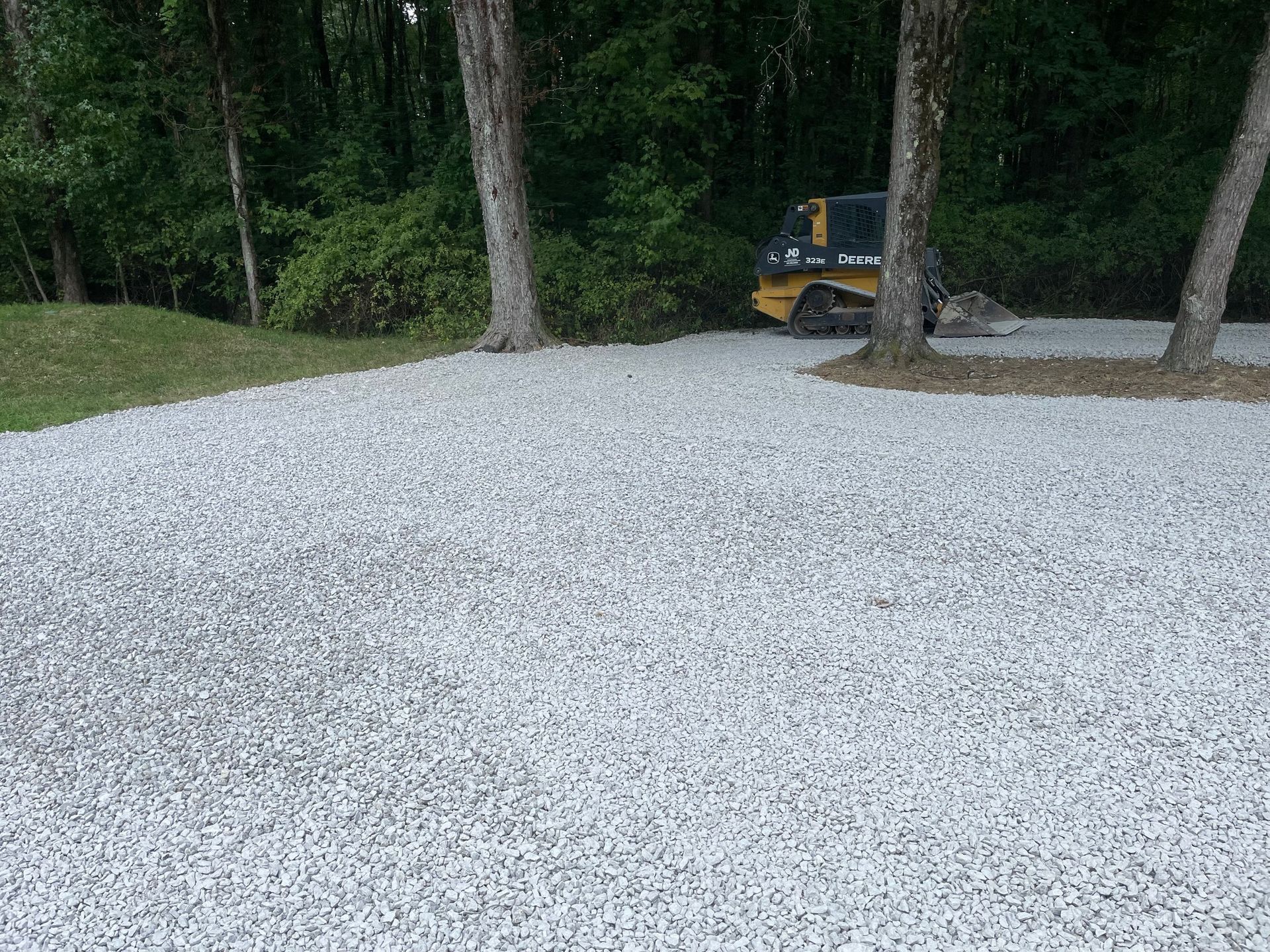 Gravel parking area beside trees and woods, with a yellow vehicle partially visible in the background