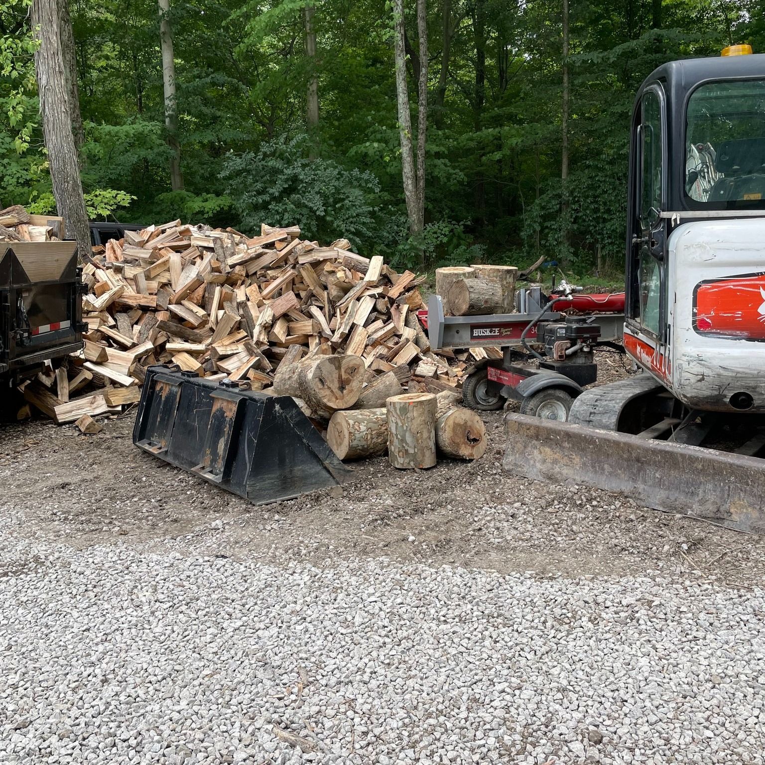 Truck loaded with firewood beside a large woodpile in a gravel lot by the woods