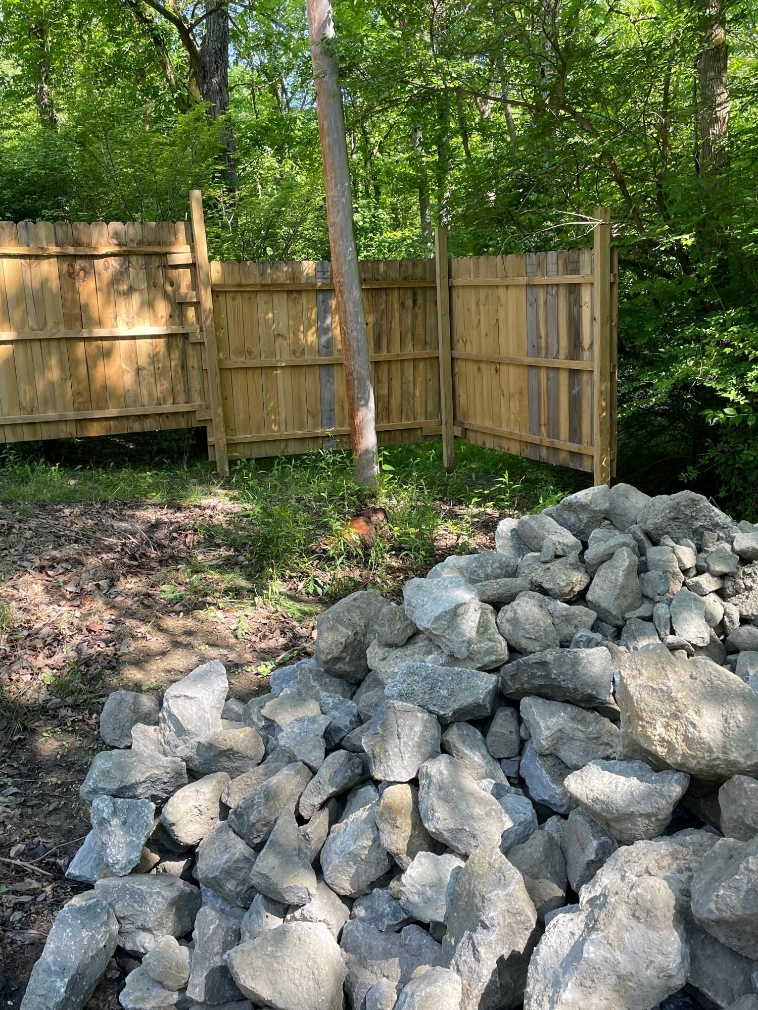 Pile of gray rocks in a yard beside a wooden fence and trees.