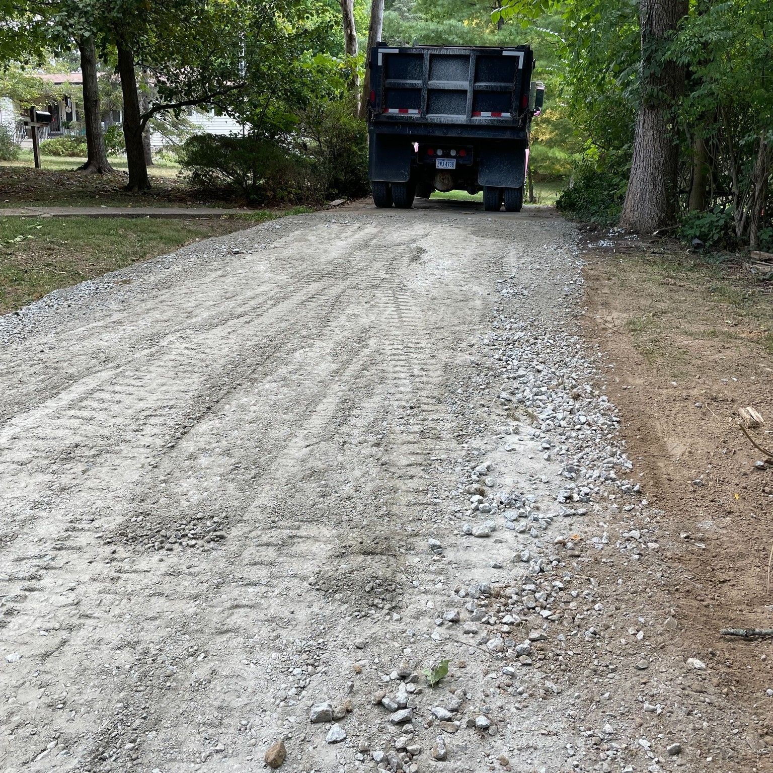 Gravel driveway with a large black dump truck parked near trees and a house on the left.