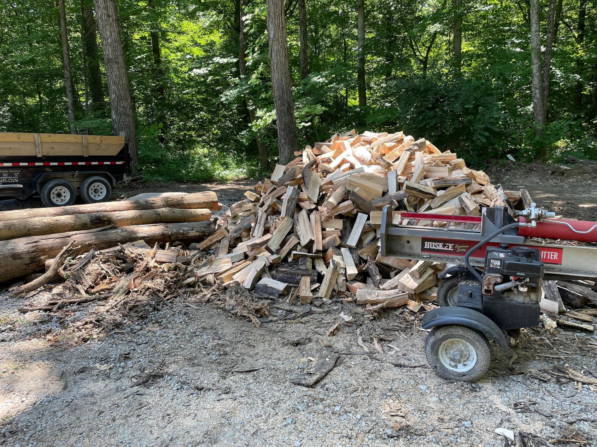 Pile of split firewood and logs beside a red log splitter in a wooded yard