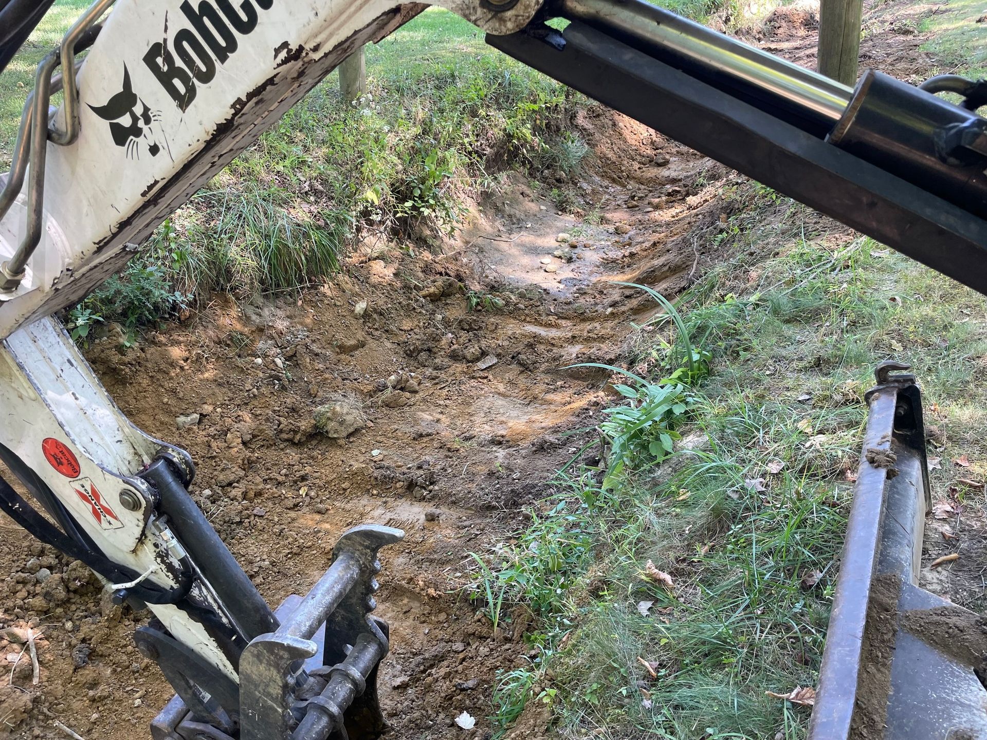 Excavator digging a narrow trench in a dirt path beside a grassy embankment.