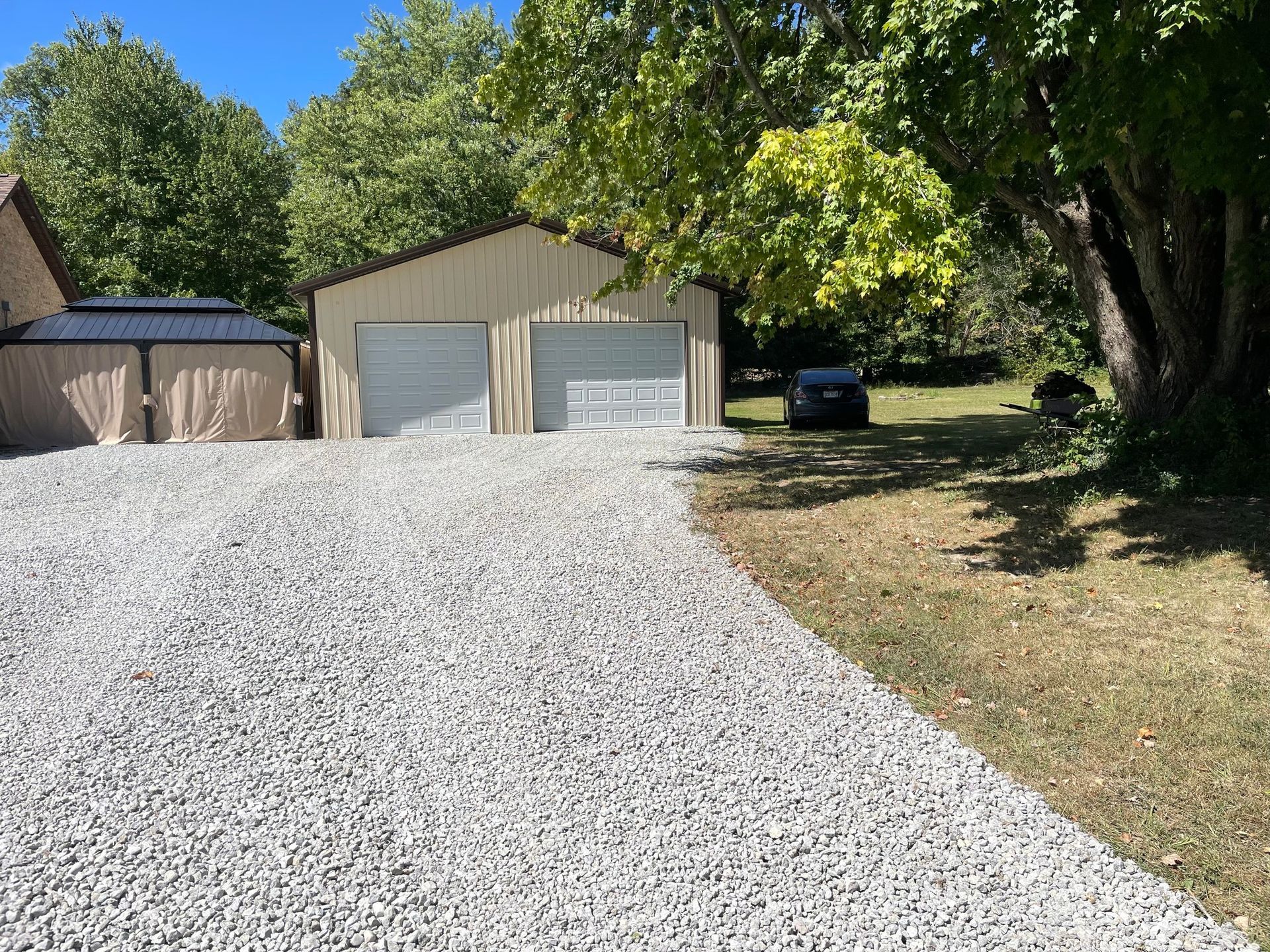 Gravel driveway leading to two white garages beside a tree-lined yard on a sunny day