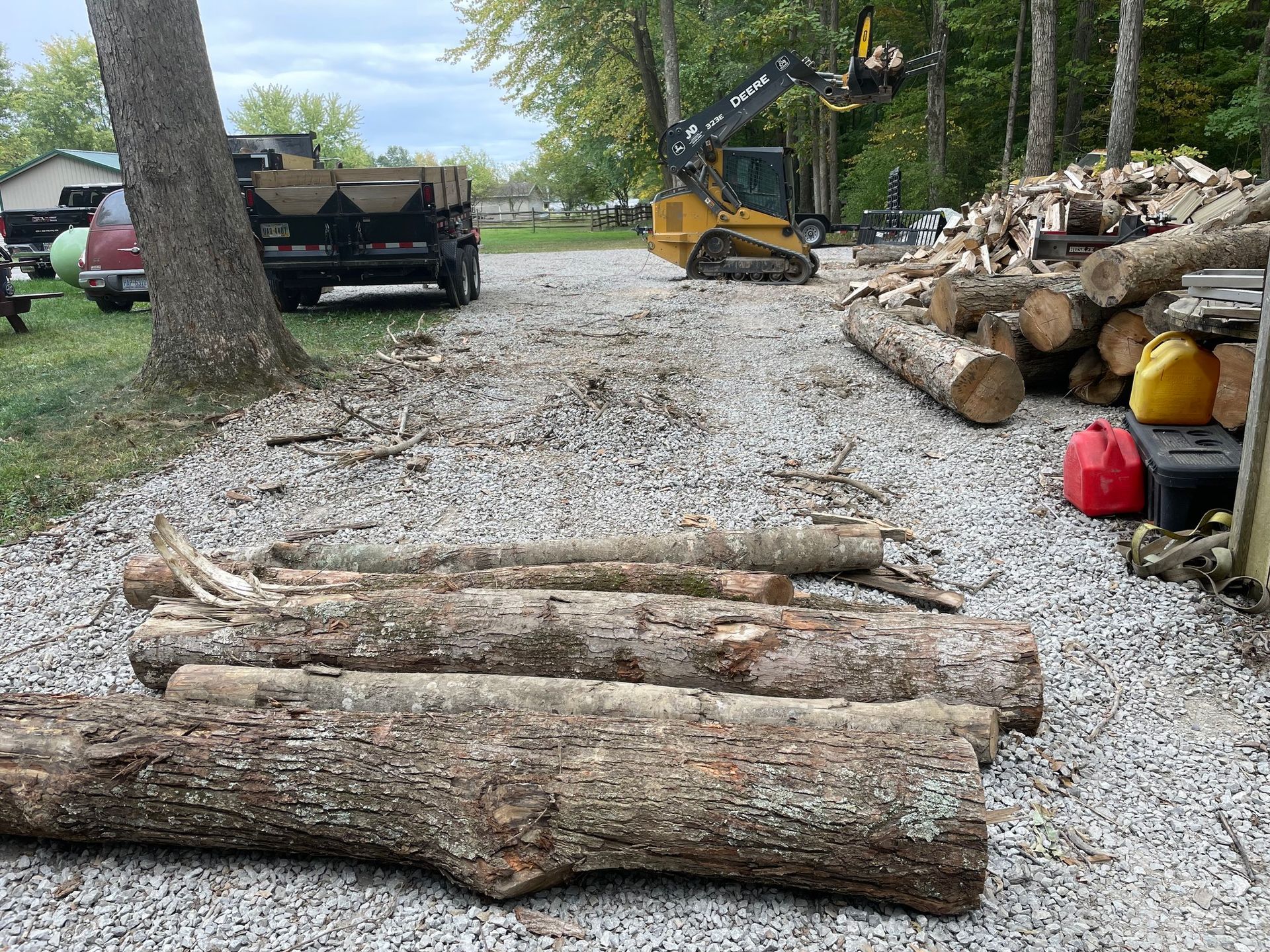 Logs stacked on gravel beside a dump truck and excavator in a wooded worksite