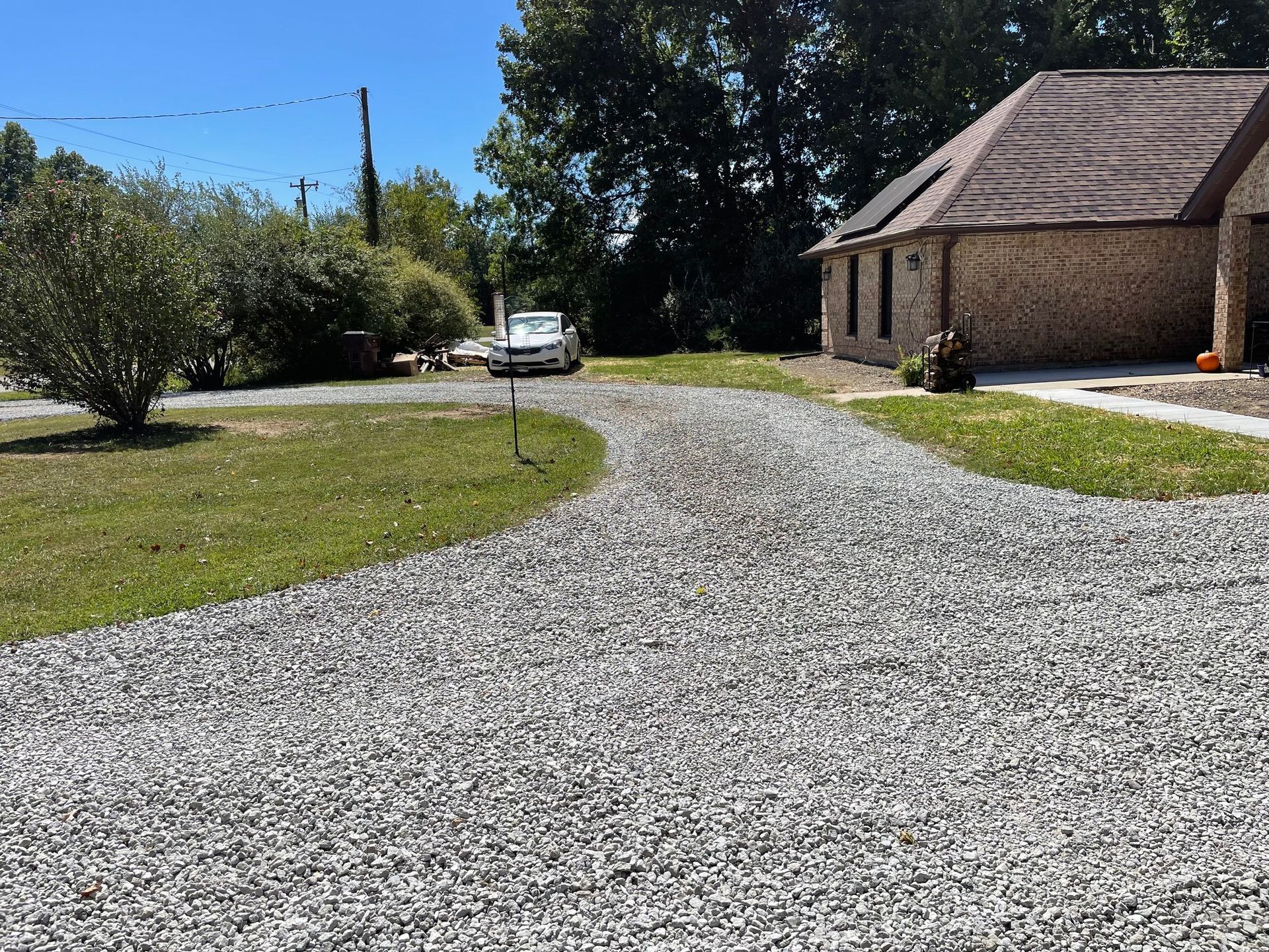 Gravel driveway curving past a brick house and trees, with a white car parked ahead.