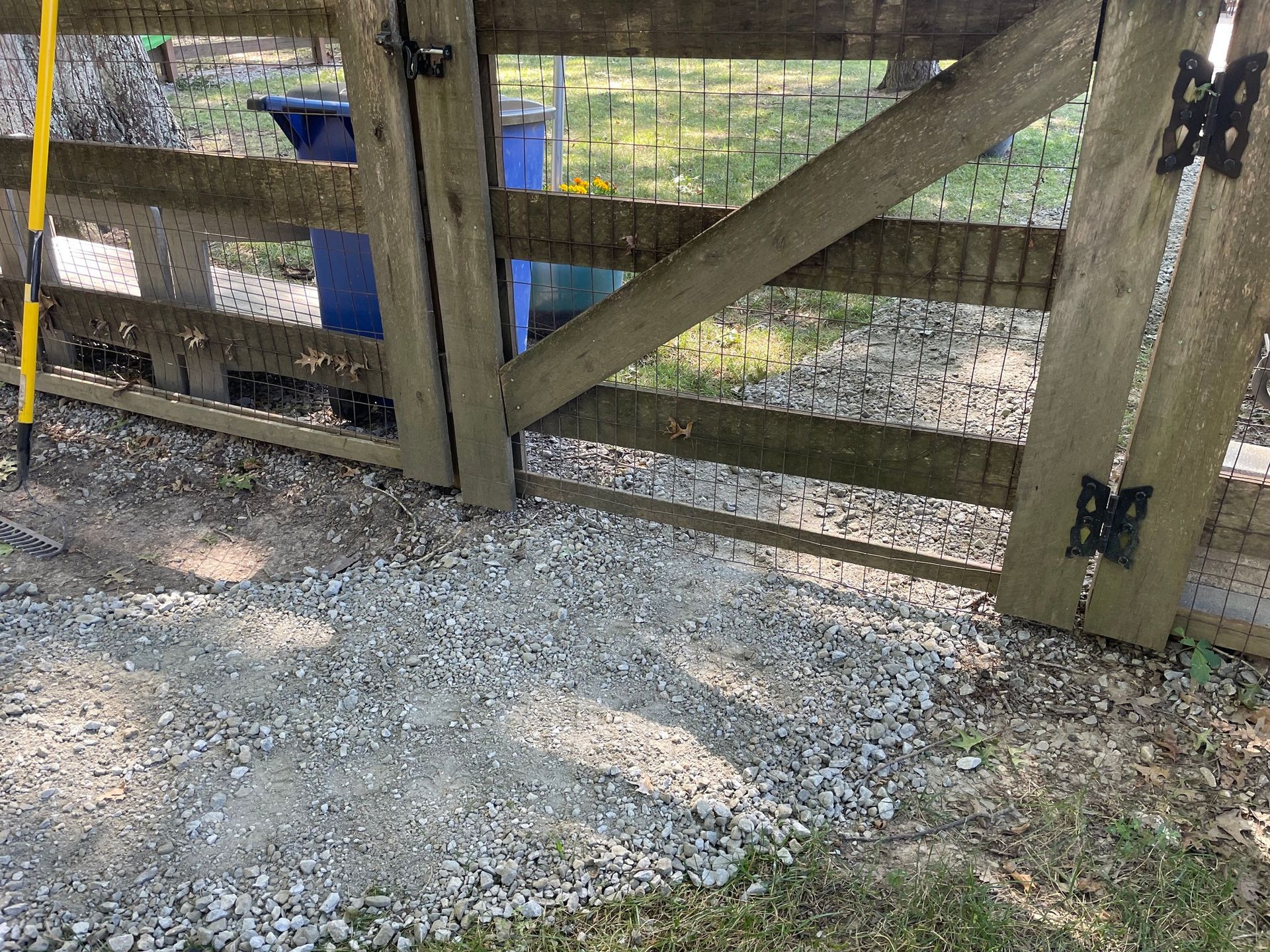 Wooden farm gate beside a gravel path, with blue buckets visible through wire fencing.