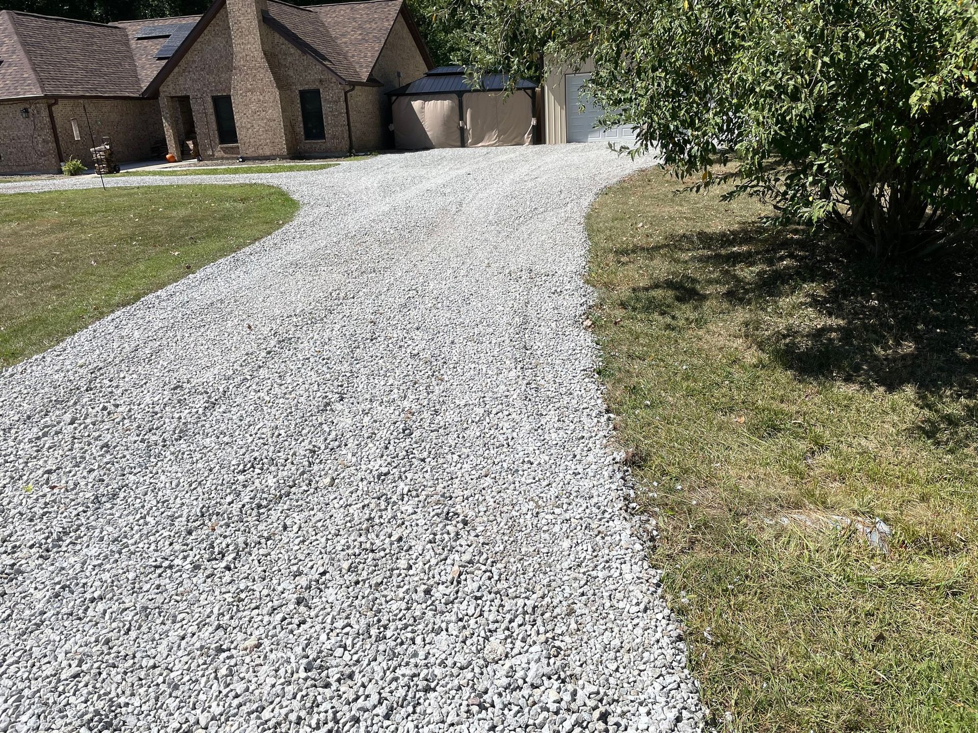 Gravel driveway curving toward a stone house beside a grassy yard and trees