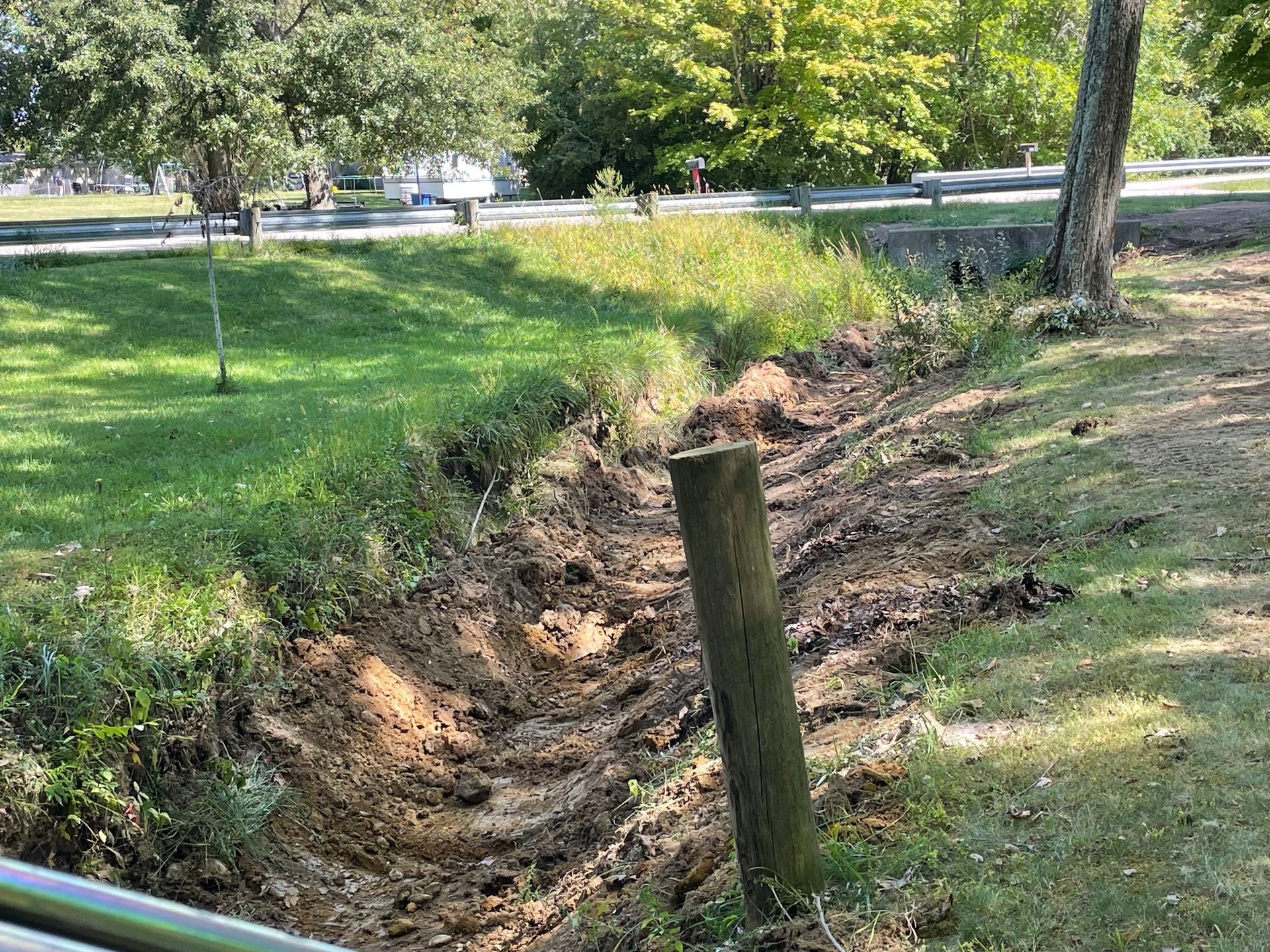 Shallow dirt trench beside a grassy park path with wooden posts and trees in the background