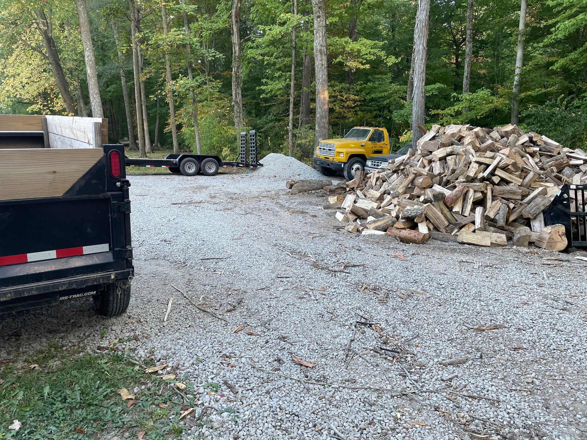 Gravel driveway with a yellow truck, black trailer, and large pile of cut logs in a wooded yard.