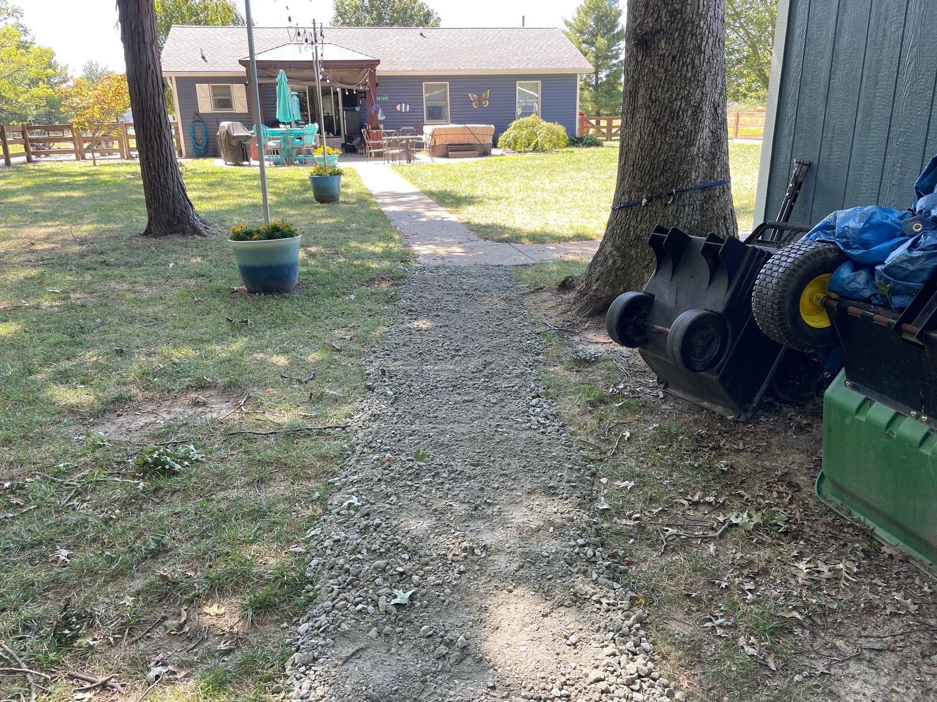 Backyard path leading to a house, with trees, potted plants, and stacked pipes on the right