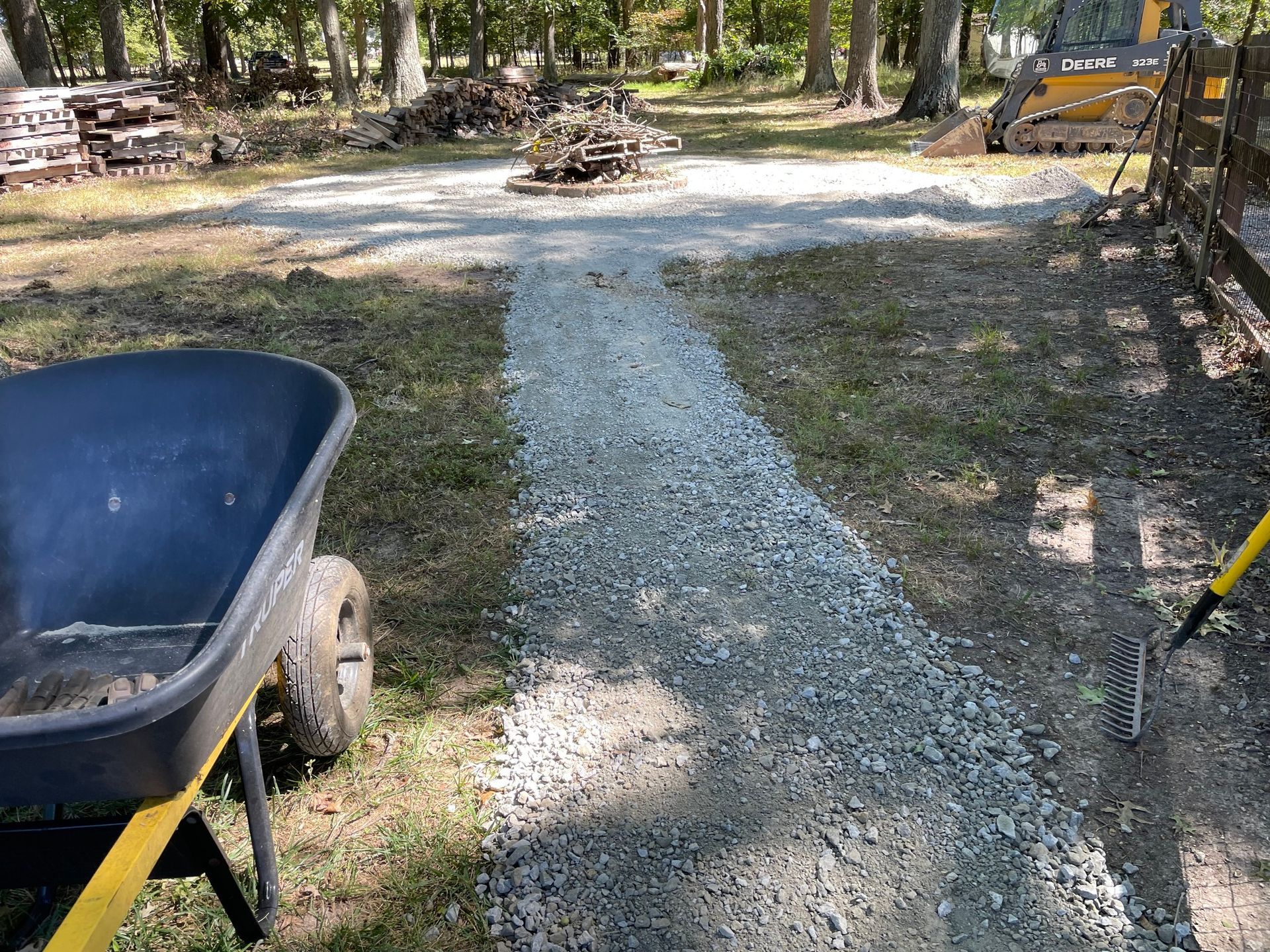 Wheelbarrow beside a gravel path leading to a small fire pit in a wooded park.
