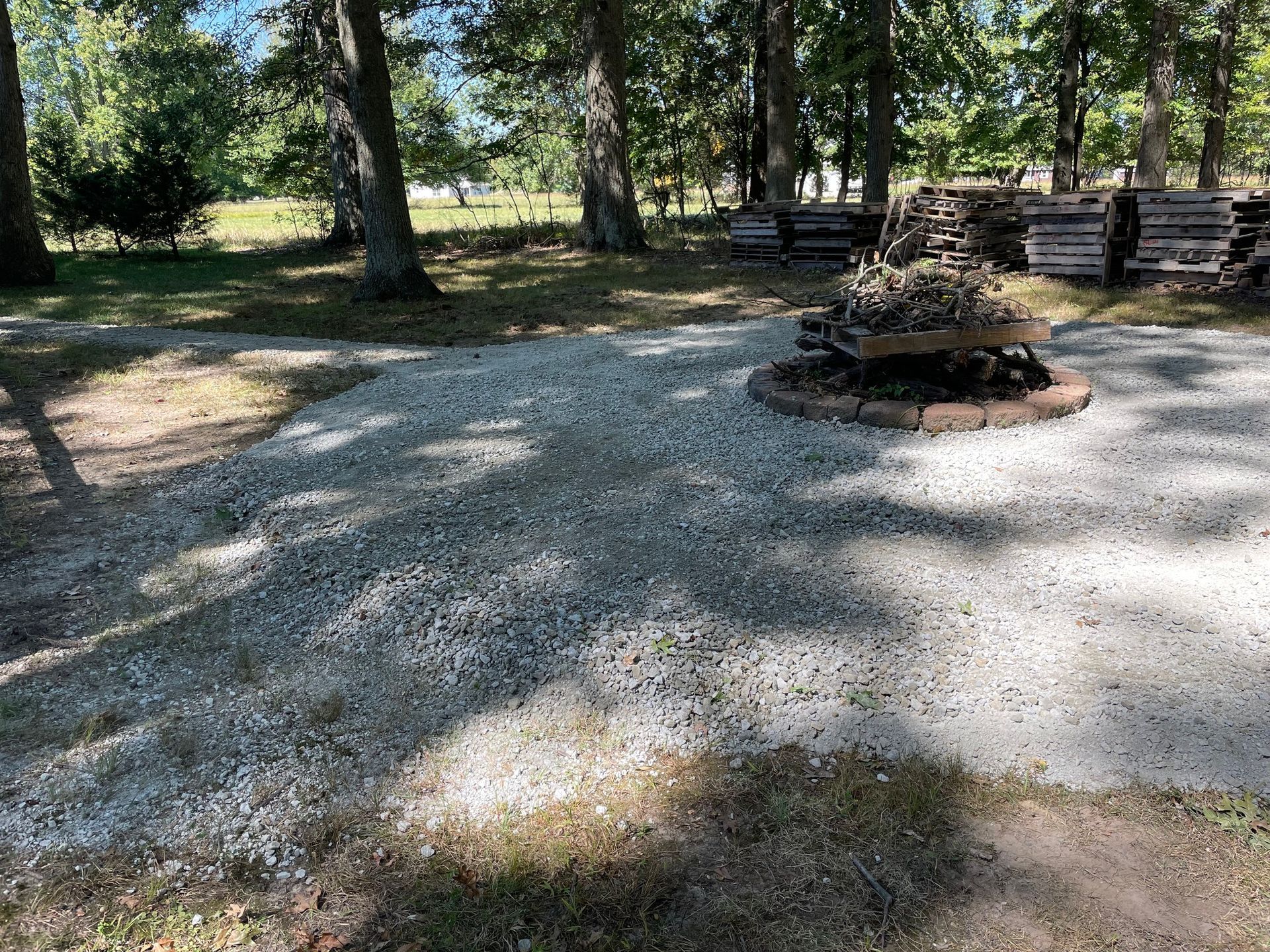 Gravel park path with trees, benches, and a small stone fire pit in a shaded clearing