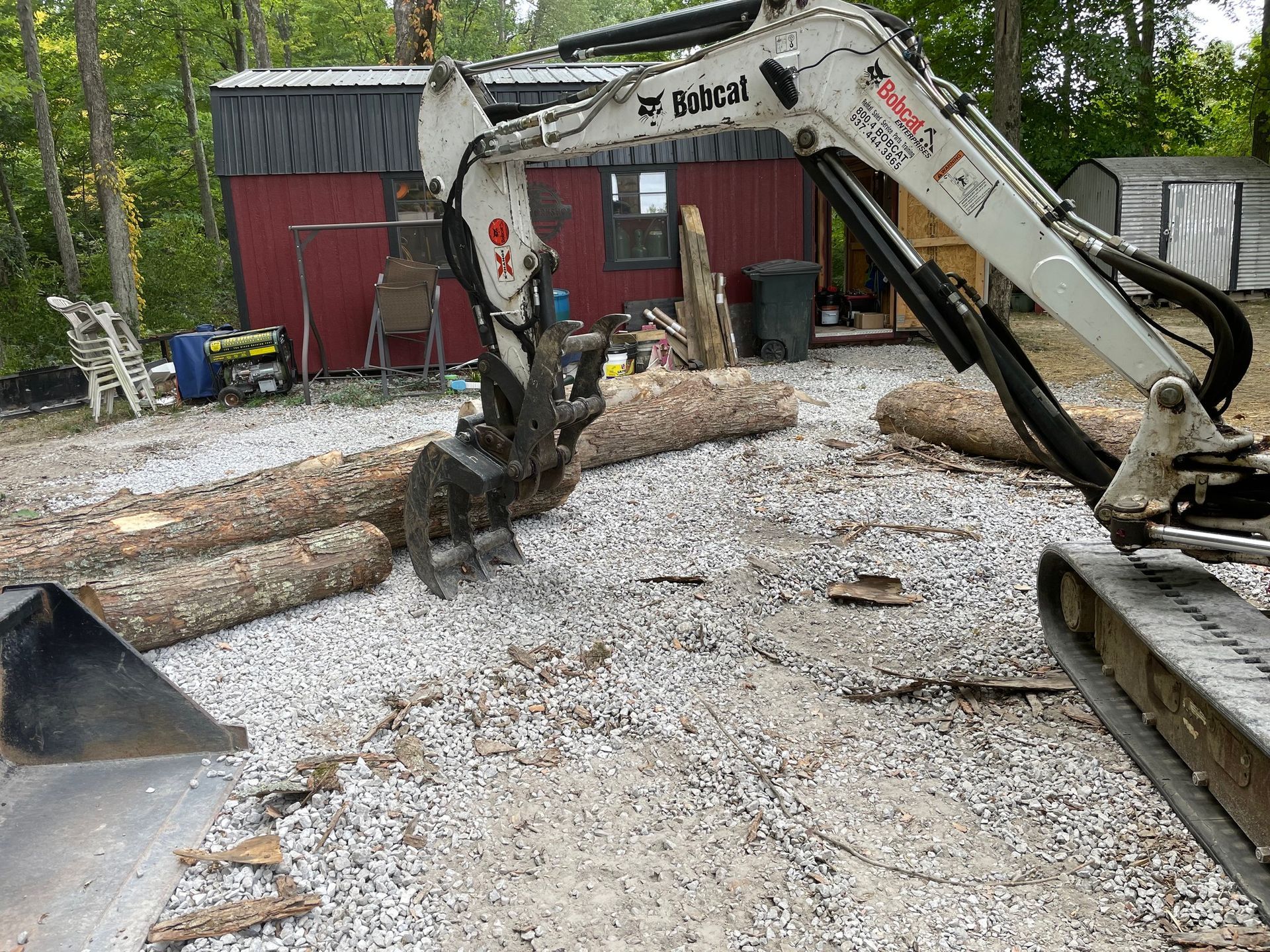 Excavator lifting logs in a gravel yard beside a red shed and stacked lumber.